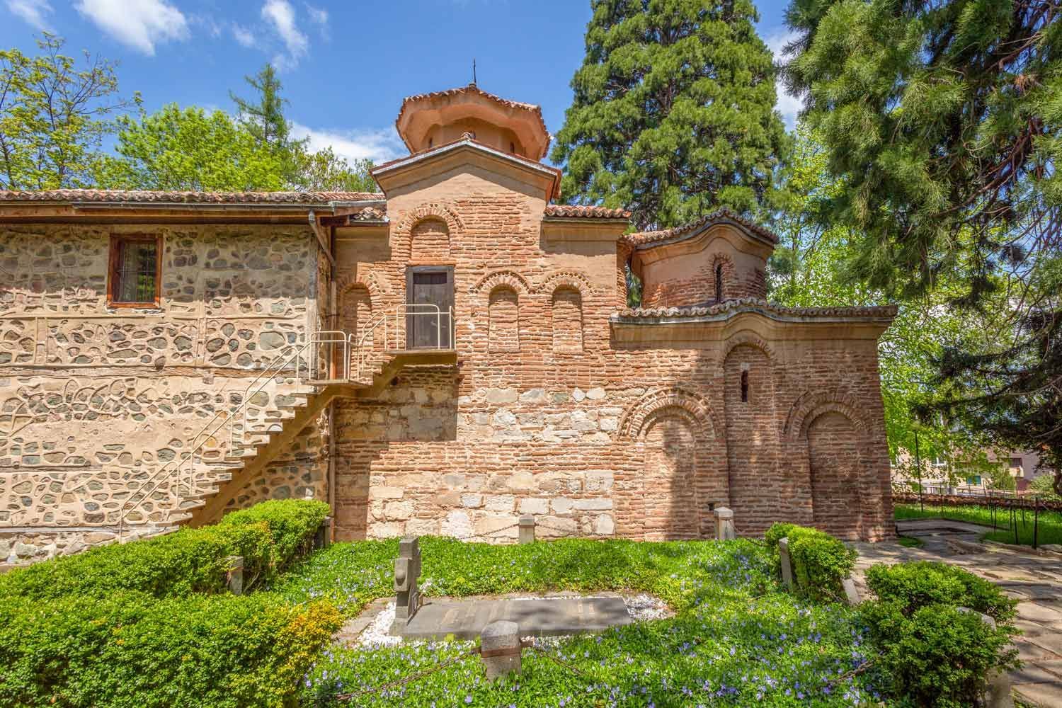Un'antica chiesa in pietra e mattoni rossi, circondata da rigogliosi alberi verdi e un piccolo giardino con una tomba.