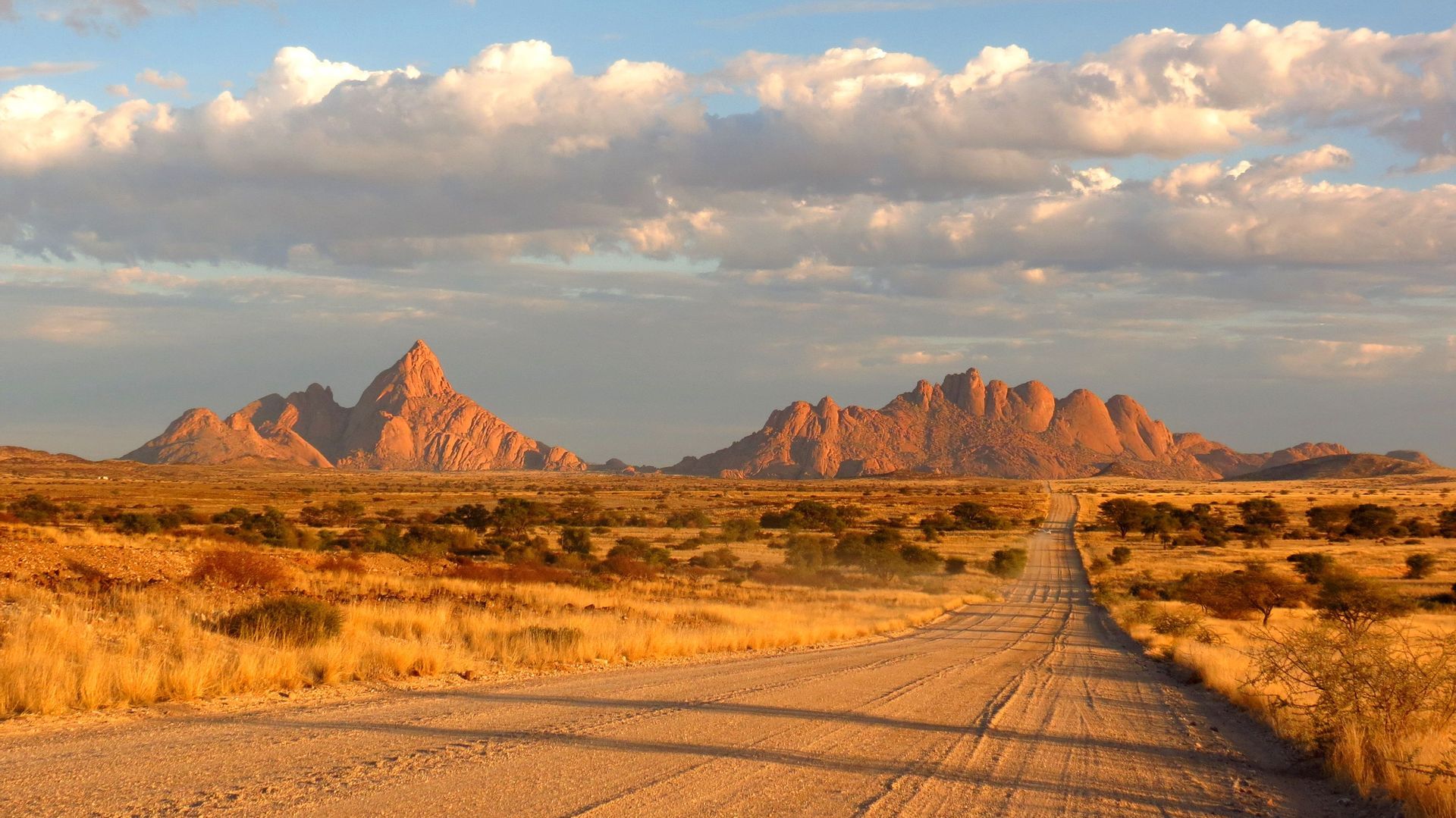 Una lunga strada sterrata si estende attraverso una secca pianura della savana verso distanti montagne rocciose illuminate dal sole sotto un cielo parzialmente nuvoloso.