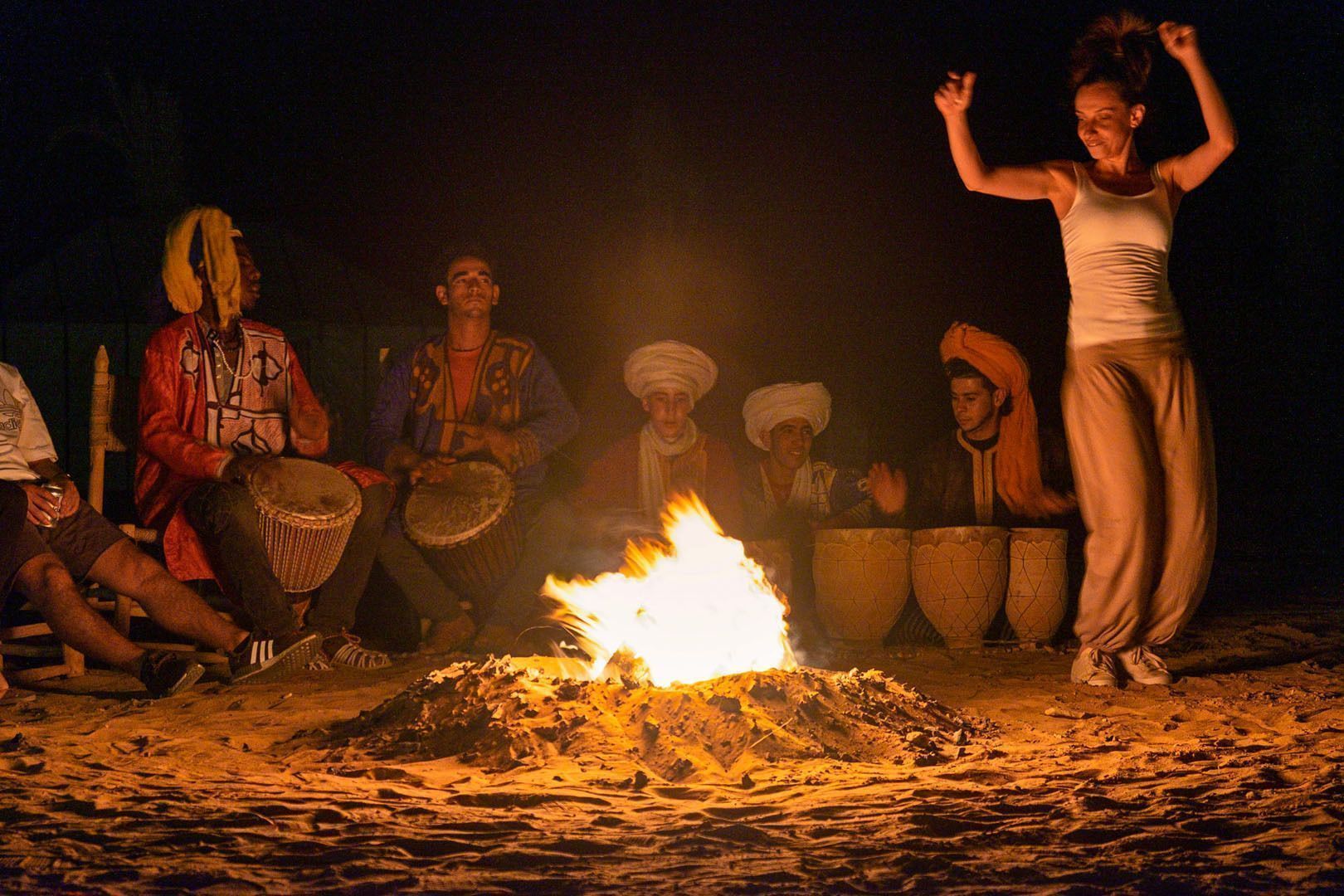A WeRoad group trip with people playing drums and dancing around a campfire on the sand at night.