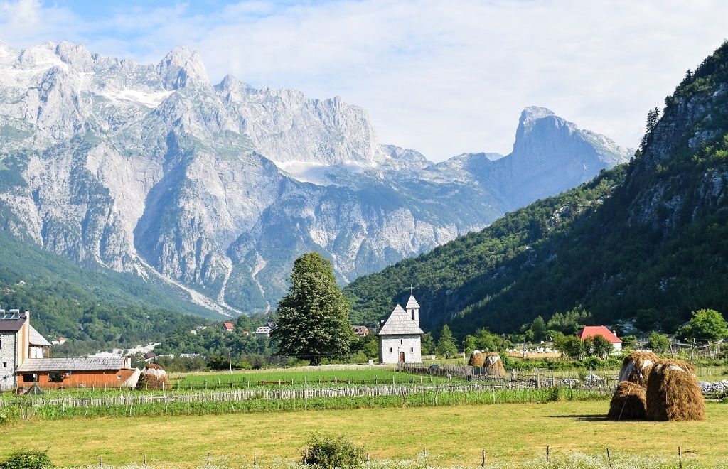 Una piccola chiesa in pietra e diversi covoni di fieno si trovano in un campo verde in una valle, con uno sfondo di grandi montagne aspre.