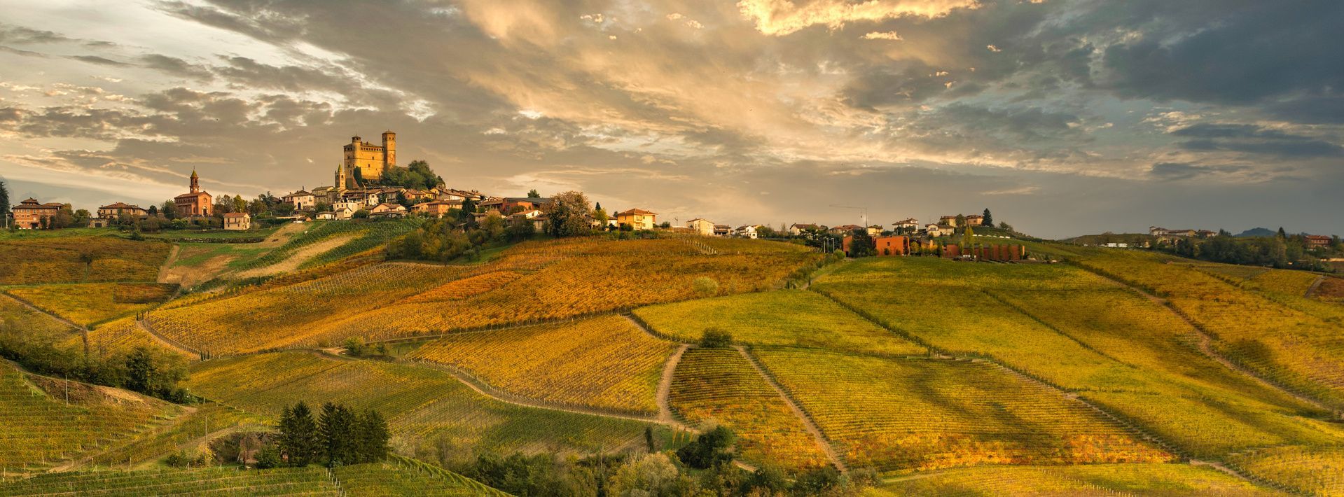 Un borgo in cima a una collina con un castello si affaccia su vigneti dorati e ondulati, sotto un cielo nuvoloso al tramonto.