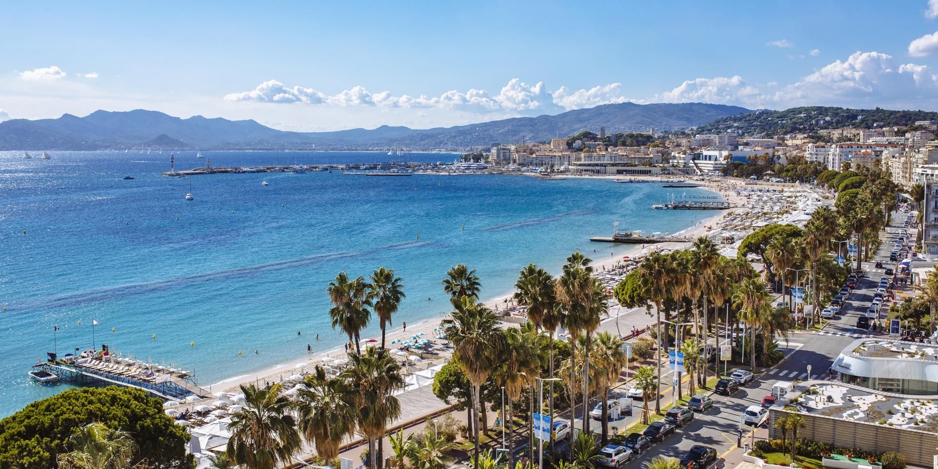 An aerial view of a coastal city with a bustling sandy beach, blue sea, and mountains in the background under a sunny sky.