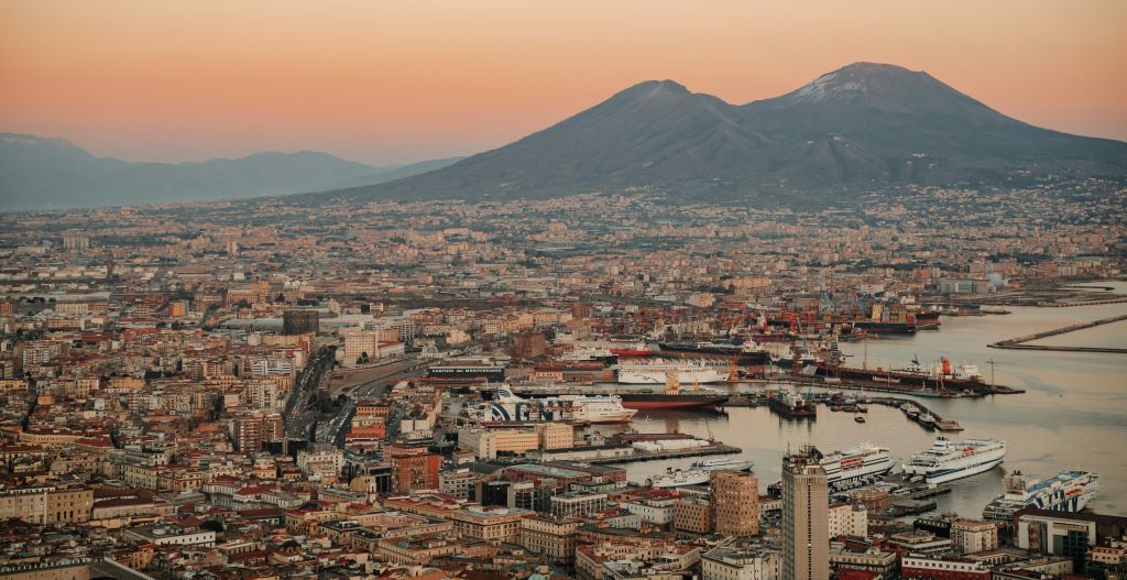 An aerial view of a sprawling coastal city and its busy port at the foot of a large volcano under a warm-toned sunset sky.