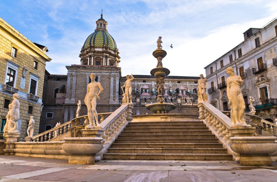 Una fontana riccamente ornata e un'ampia scalinata fiancheggiata da statue classiche nude in una piazza storica sotto un cielo azzurro.