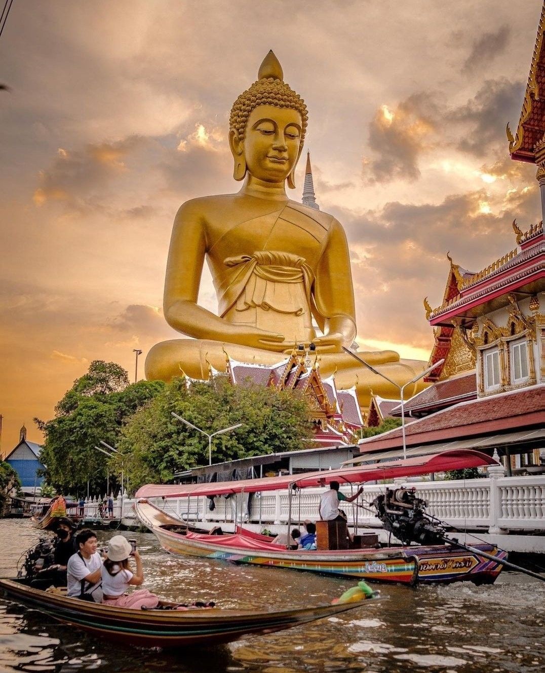 Des bateaux à longue queue naviguent sur un canal, avec une statue géante de Bouddha doré et un temple en arrière-plan, au coucher du soleil.