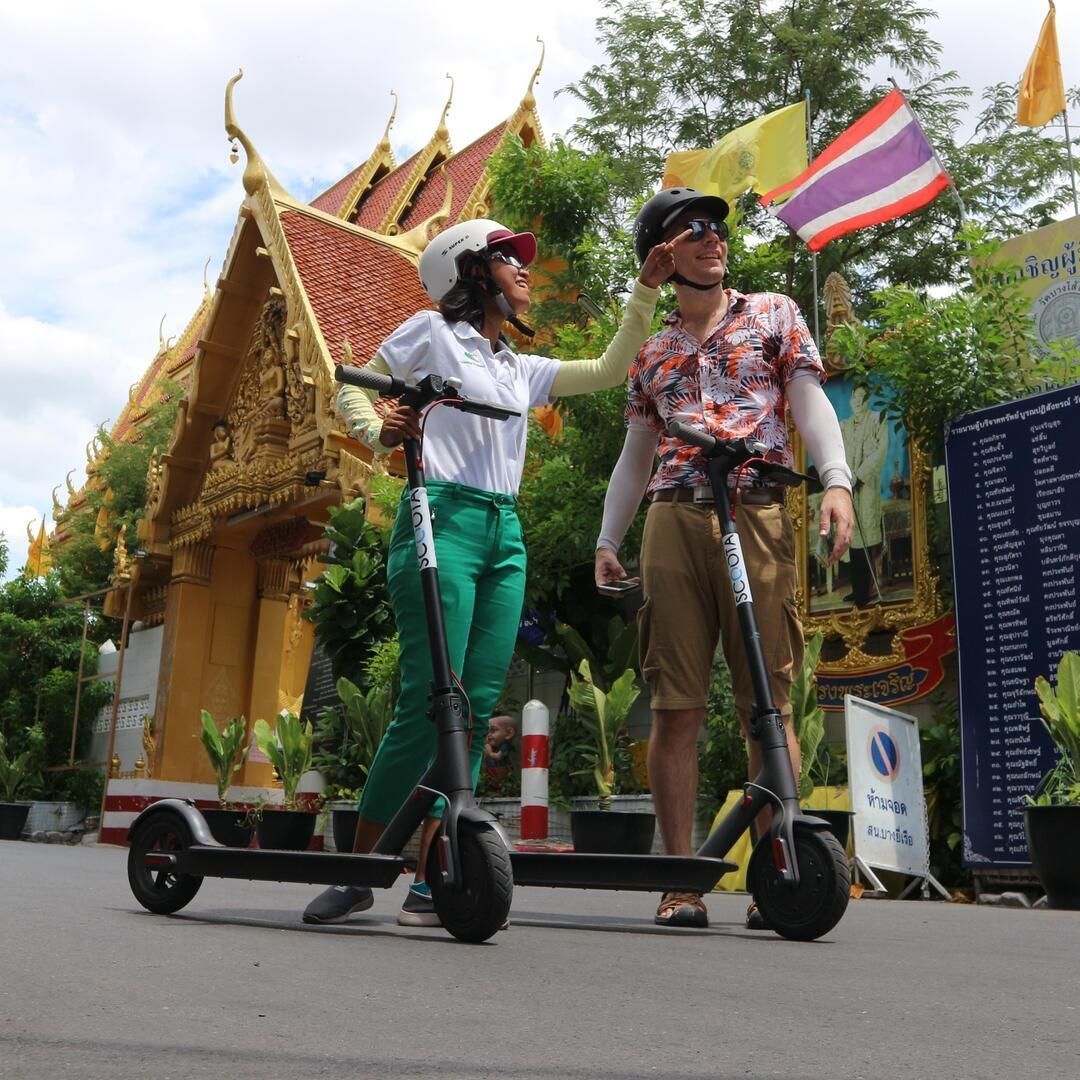 Une femme ajuste le casque d'un homme alors qu'ils se tiennent avec des trottinettes électriques devant un temple doré orné d'un toit de tuiles rouges.