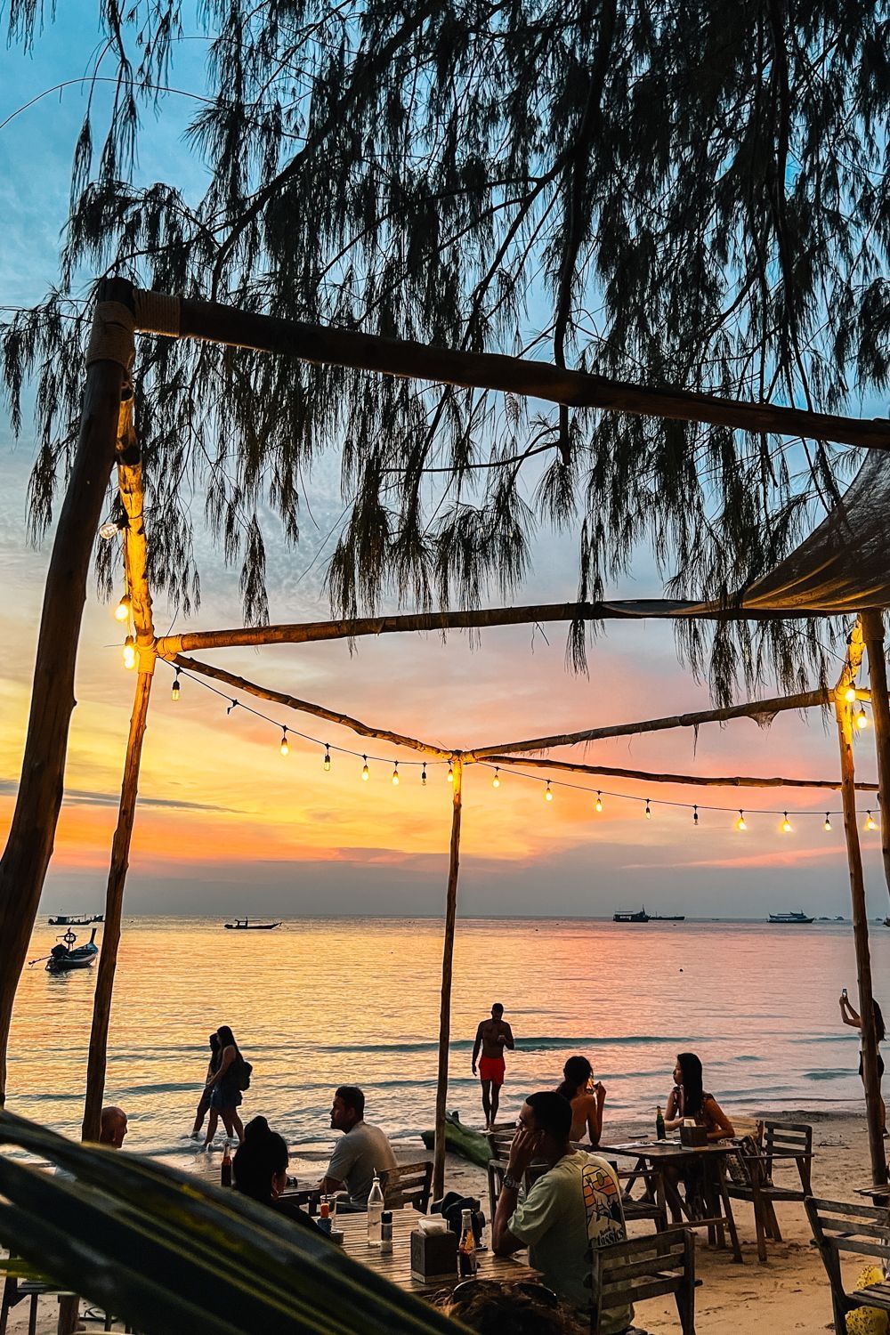 Un groupe WeRoad se détend dans un bar de plage sous des guirlandes lumineuses, regardant le coucher de soleil sur la mer.