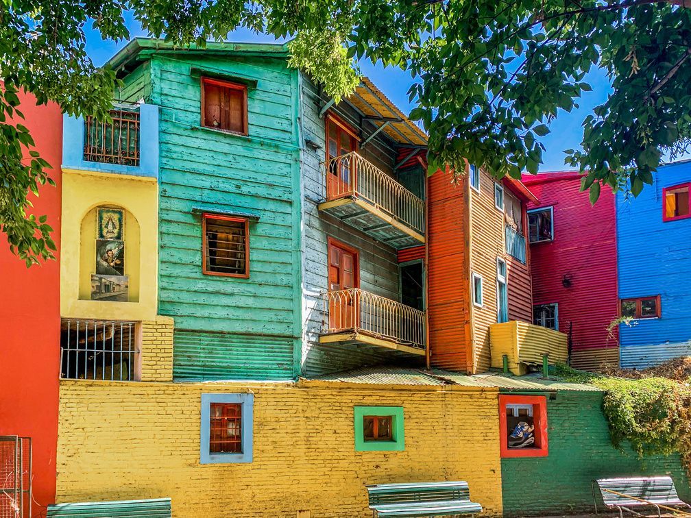 Exterior view of stacked, colorful buildings, painted turquoise, yellow, red, and blue, under green tree leaves and a clear sky.