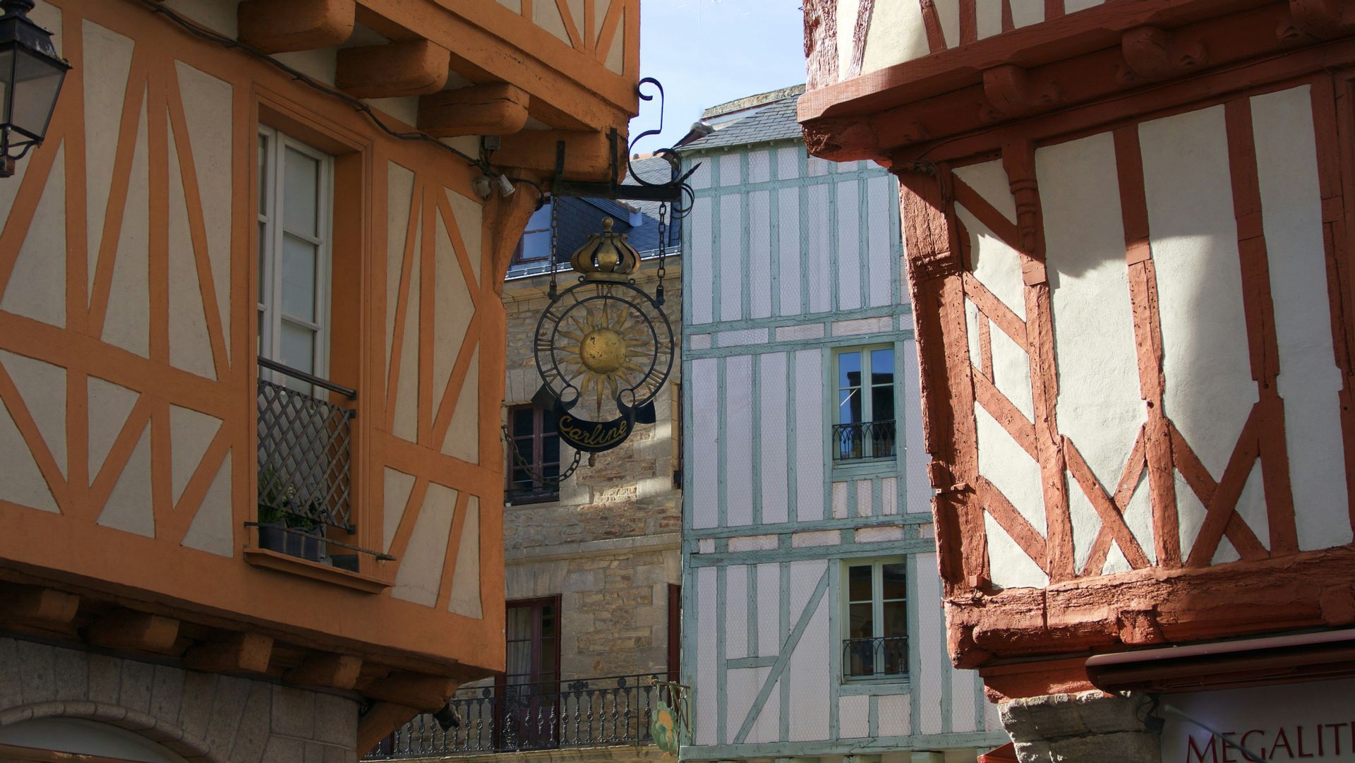Des maisons à colombages colorées avec poutres apparentes bordent une rue étroite, avec une enseigne métallique ornée suspendue au-dessus.