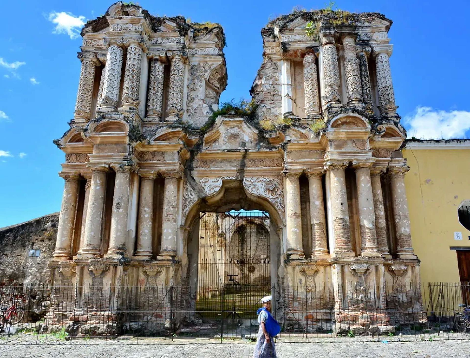 A person in traditional clothing walks on a cobblestone street in front of a large, ornate, and crumbling stone facade.