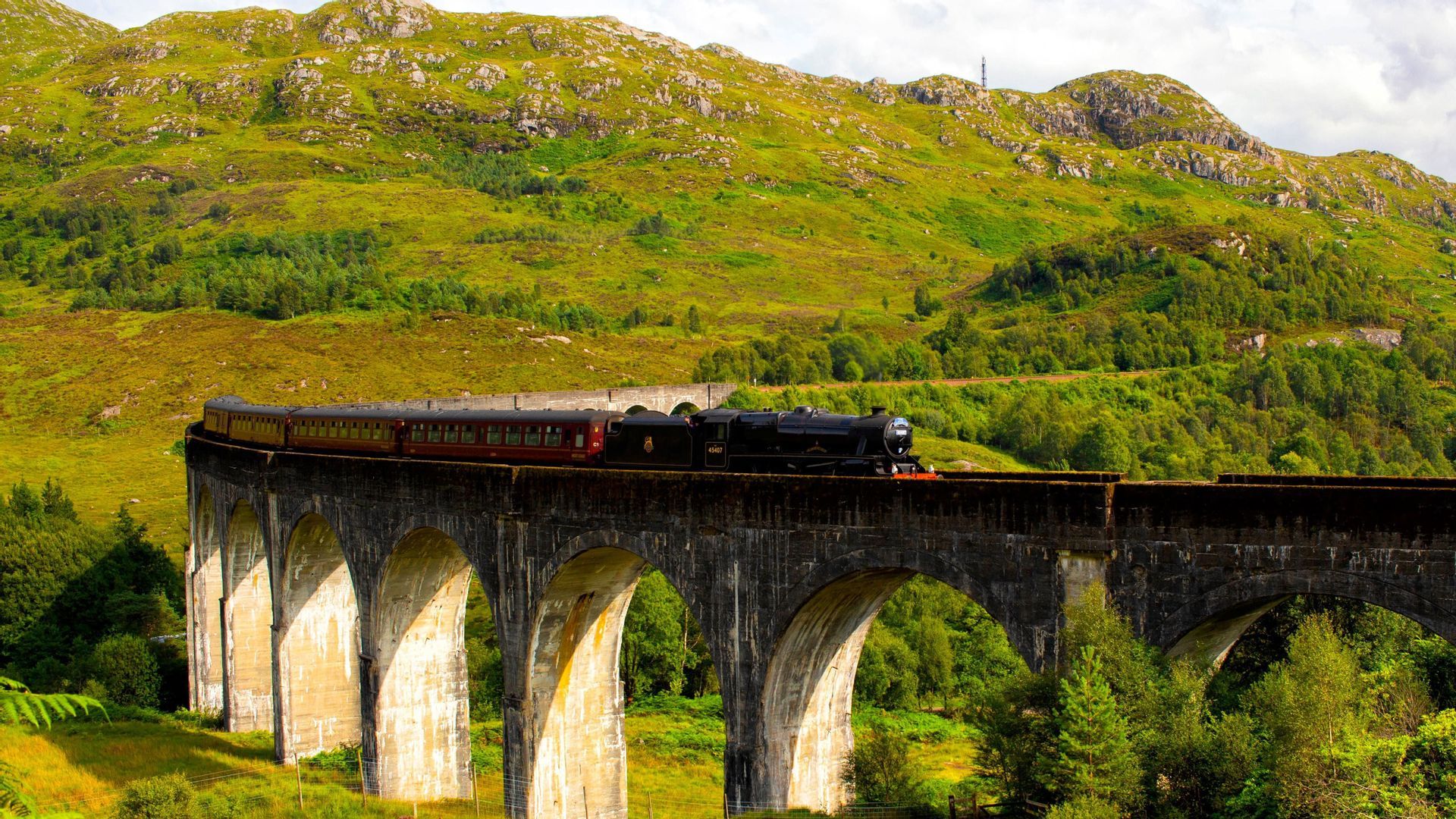 Un treno a vapore nero con carrozze rosse attraversa un viadotto in pietra, con una rigogliosa montagna verde sullo sfondo.