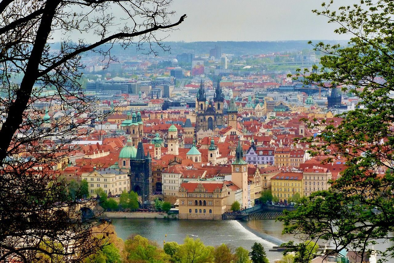 Vue plongeante d'une ville européenne historique avec des toits de tuiles rouges et des flèches, encadrée par des branches d'arbres avec une rivière en contrebas.