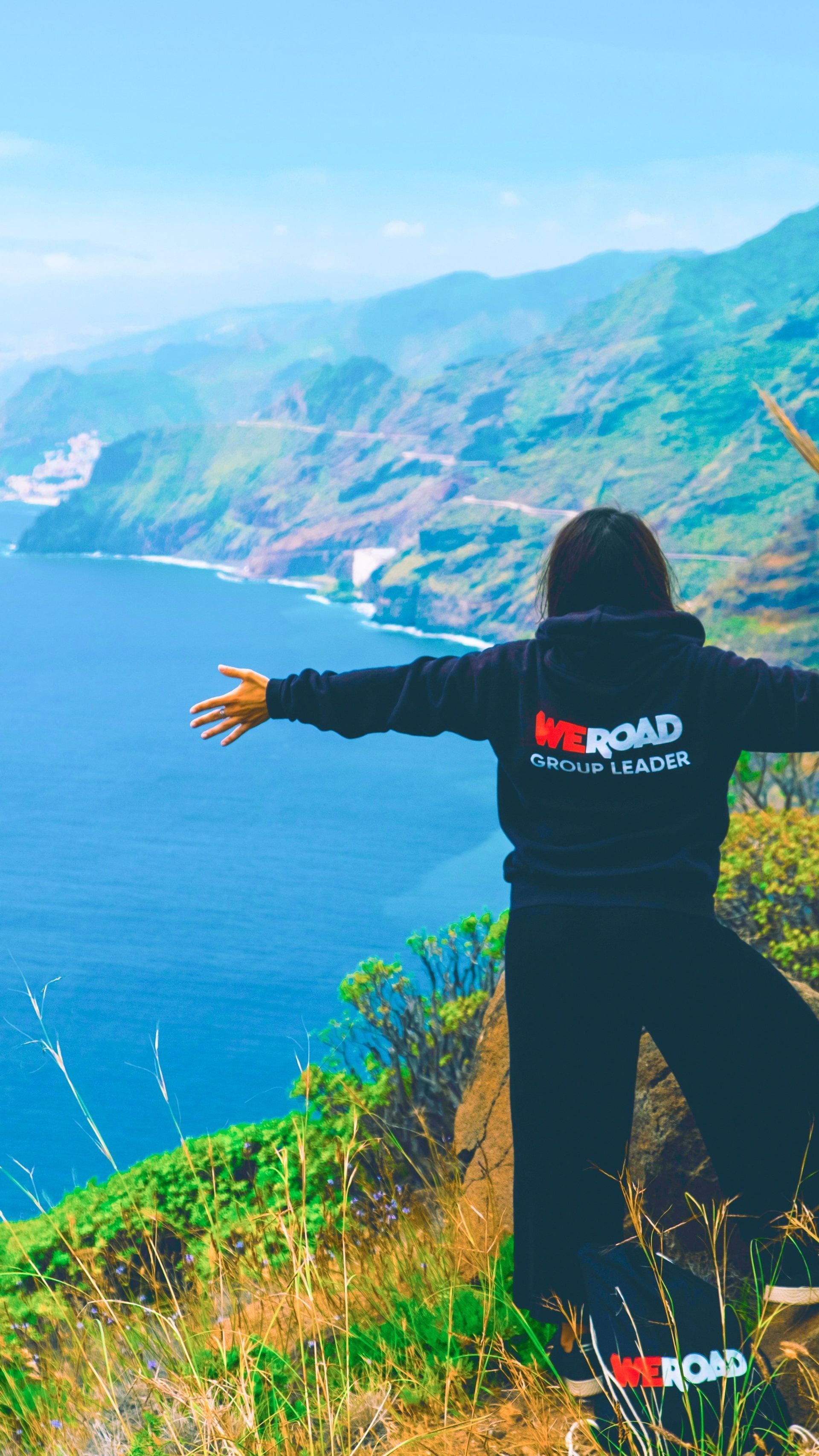 Un capo gruppo WeRoad si erge su una scogliera erbosa con le braccia aperte, ammirando una costa frastagliata e un vasto oceano blu.