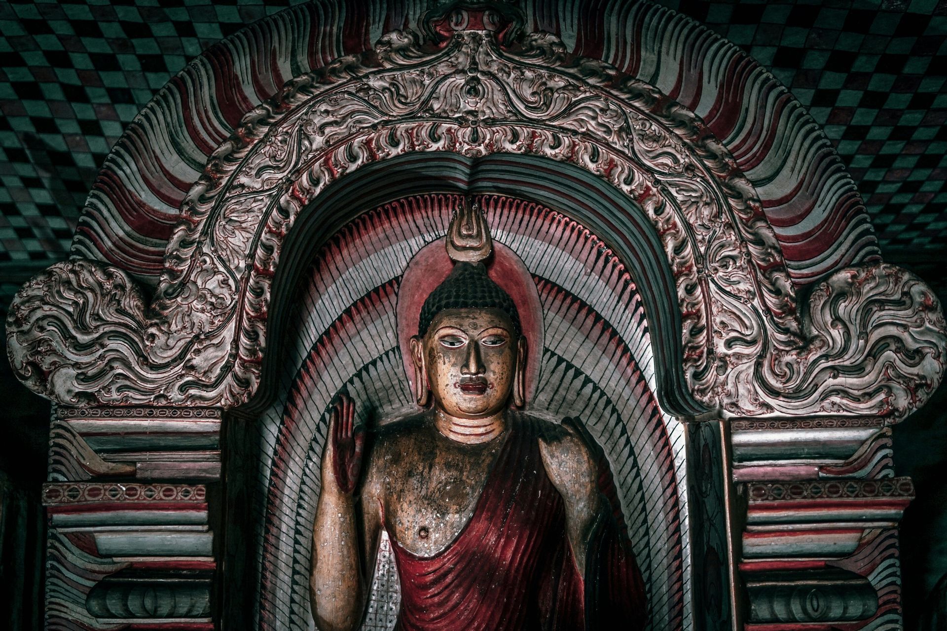 Una statua dipinta di Buddha con la mano alzata, collocata in una nicchia ad arco riccamente decorata.