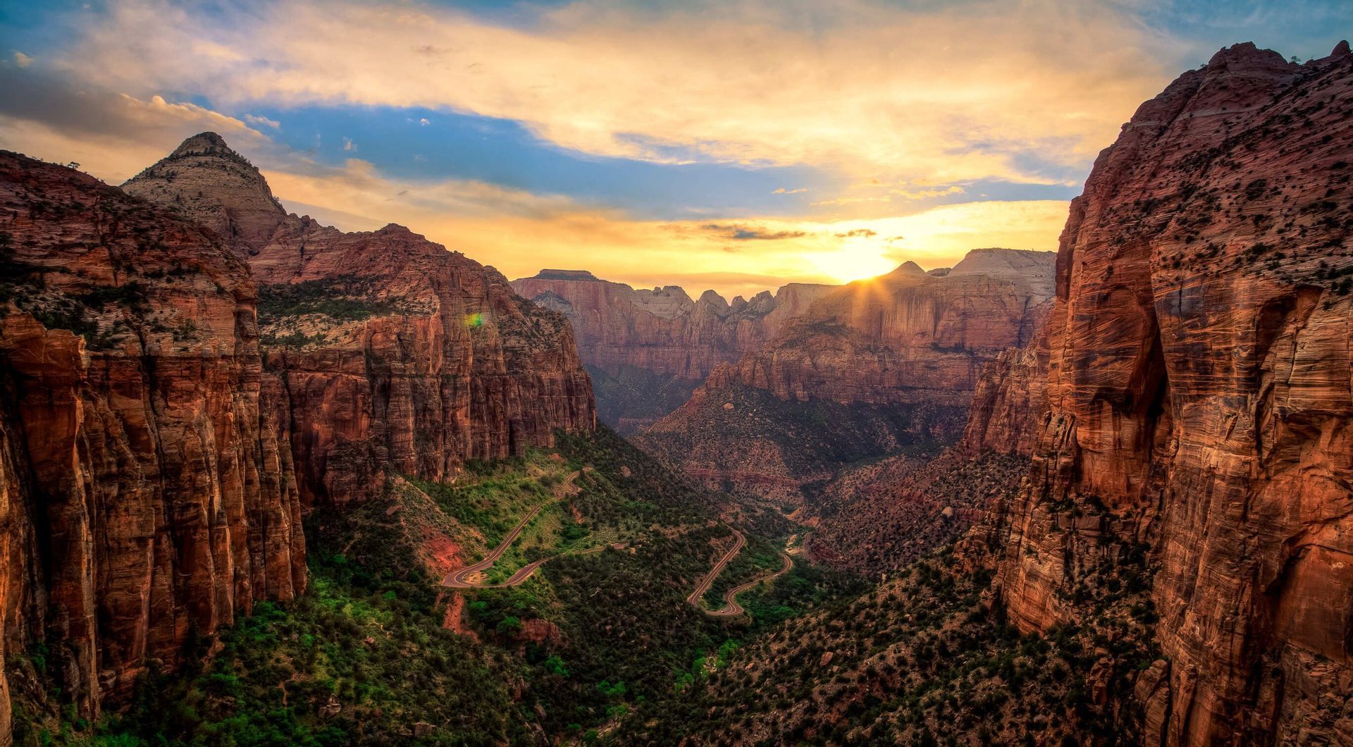 Il sole tramonta su un canyon profondo, illuminando le rosse pareti rocciose e una strada tortuosa nella valle verde sottostante.