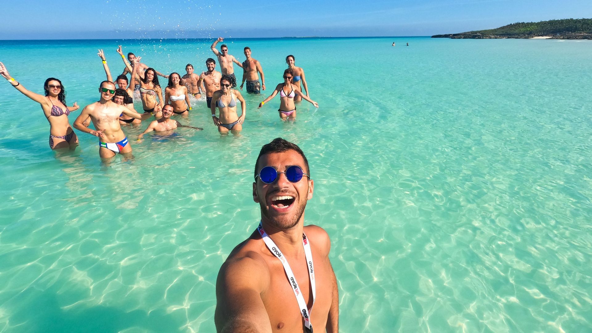 Ein Mann macht ein Selfie mit einer WeRoad-Gruppe, während sie an einem sonnigen Tag gemeinsam im klaren türkisfarbenen Meer posieren.