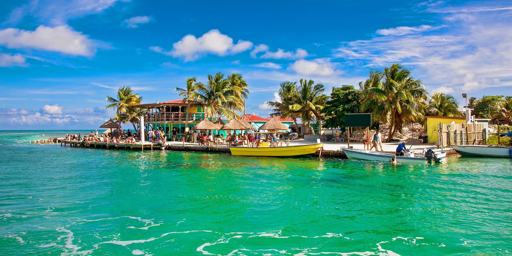 A WeRoad group trip relaxes on a wooden pier by a tropical bar, with boats docked in the turquoise water and palm trees under a blue sky.
