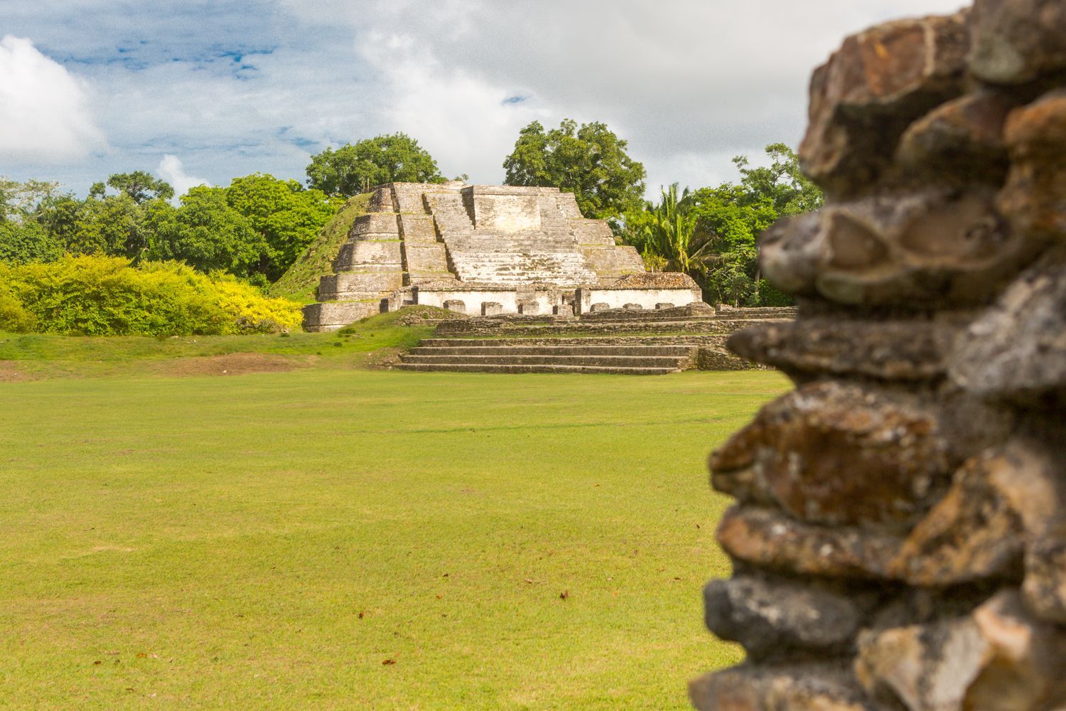 Une grande pyramide de pierre à degrés se dresse dans une clairière verdoyante, entourée d'arbres luxuriants sous un ciel nuageux.