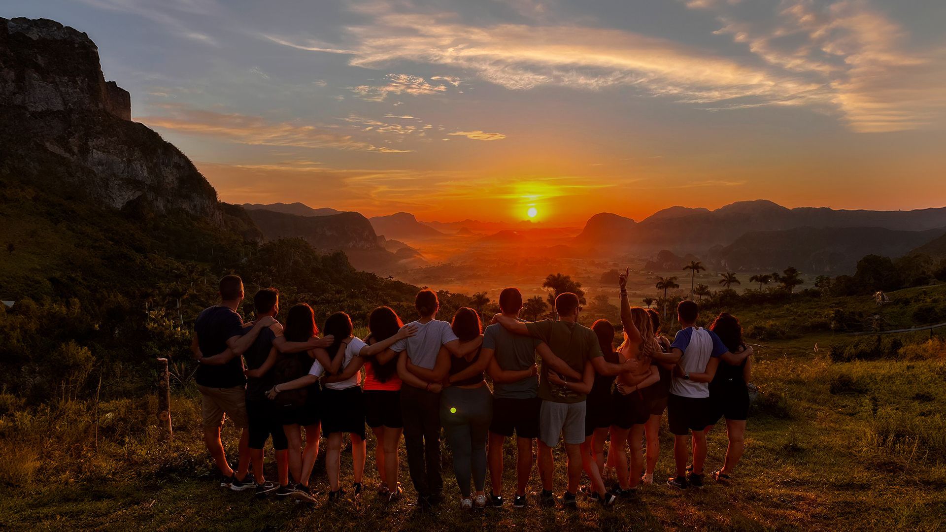 Un gruppo WeRoad in viaggio, abbracciato, che ammira il tramonto su una valle di montagna da un punto panoramico.