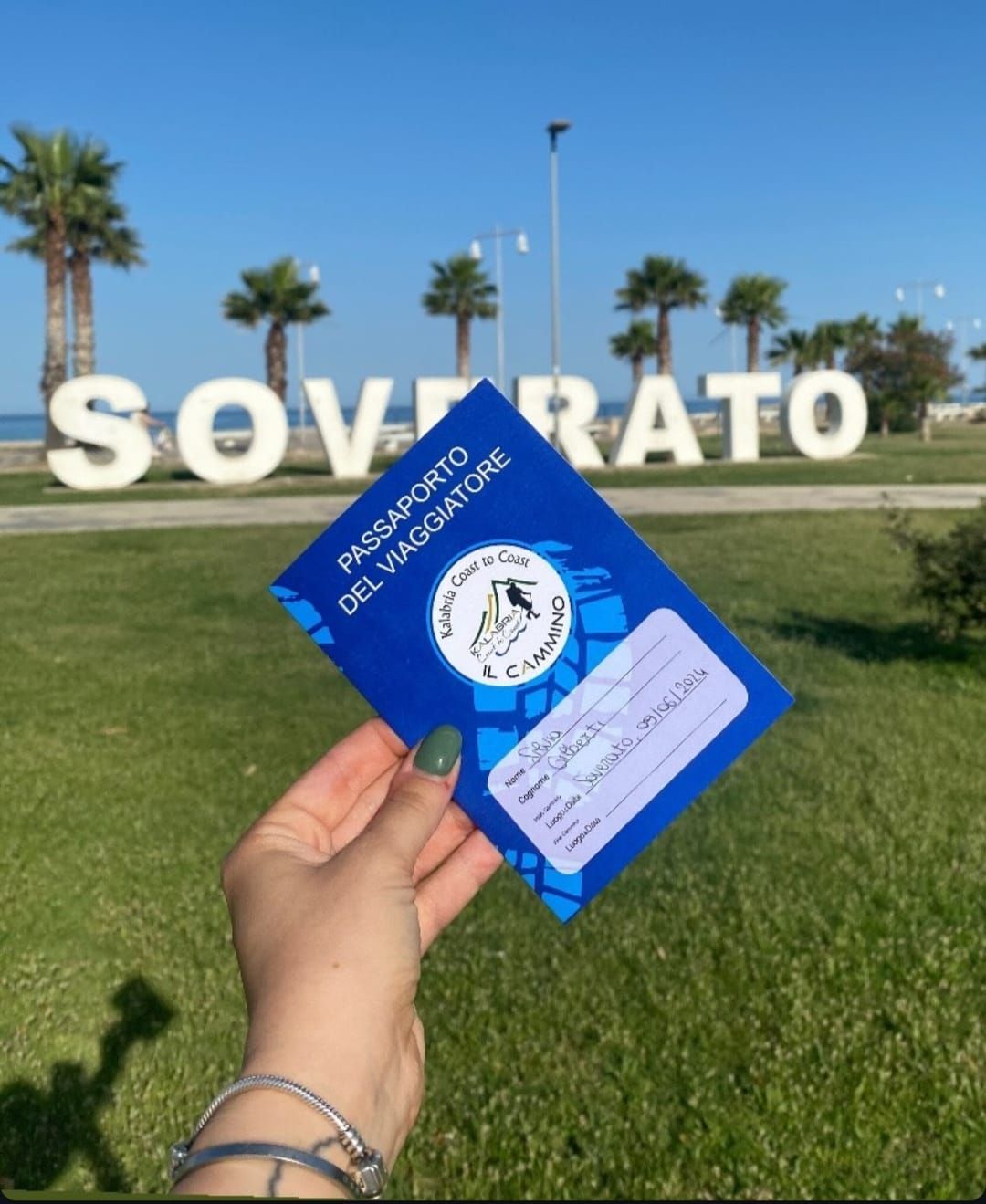 Una mano regge un 'Passaporto del Viaggiatore' davanti a un grande cartello 'SOVERATO' su un prato con palme.