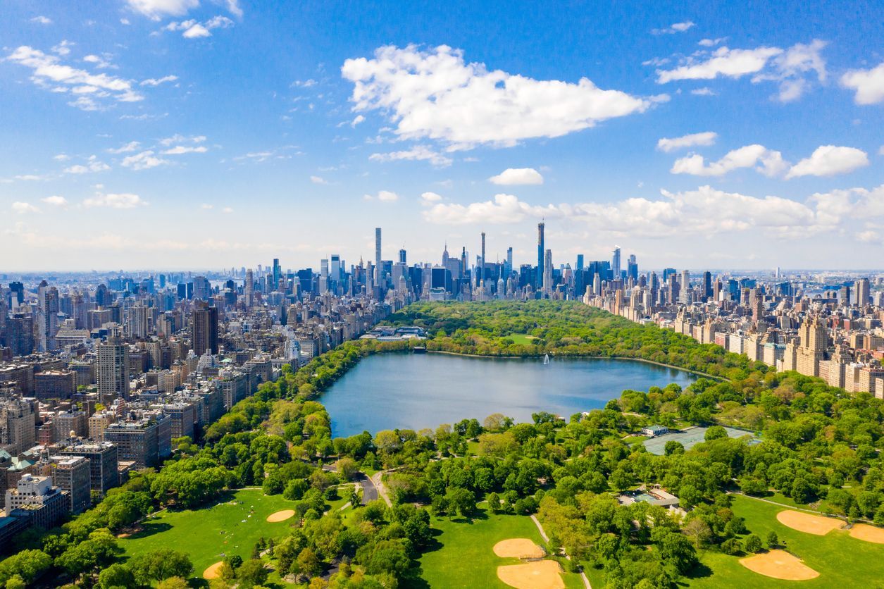 Vista aerea di un grande parco urbano con un lago centrale, delimitato da una fitta distesa di grattacieli sotto un cielo azzurro con nuvole.