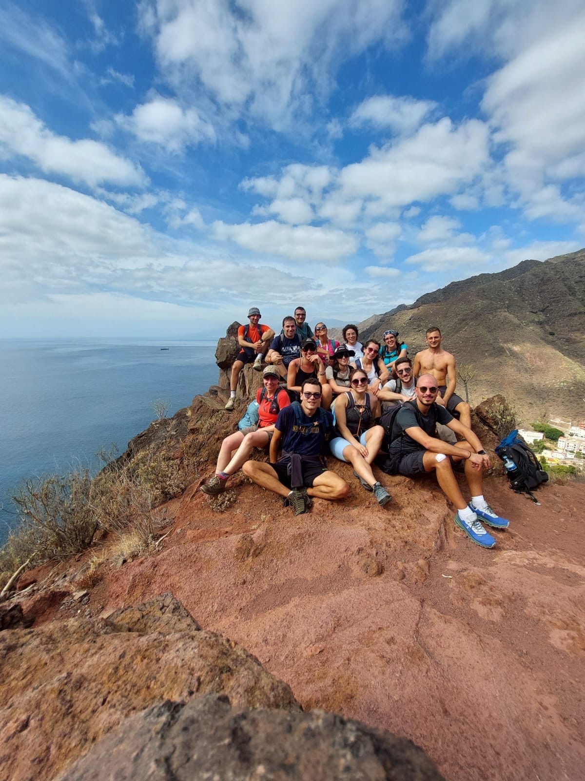 Un gruppo WeRoad in viaggio posa per una foto su una scogliera rocciosa che si affaccia sul mare e sulle montagne sotto un cielo azzurro.