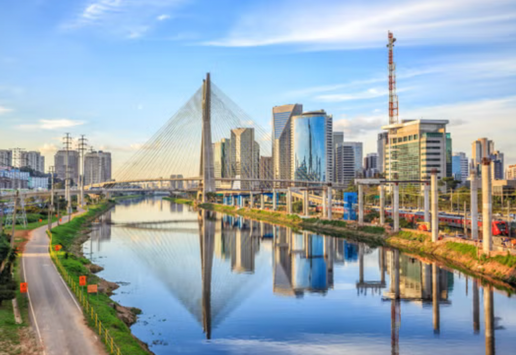 A cable-stayed bridge spans a river in a modern city, with skyscrapers reflected in the calm water under a blue sky.