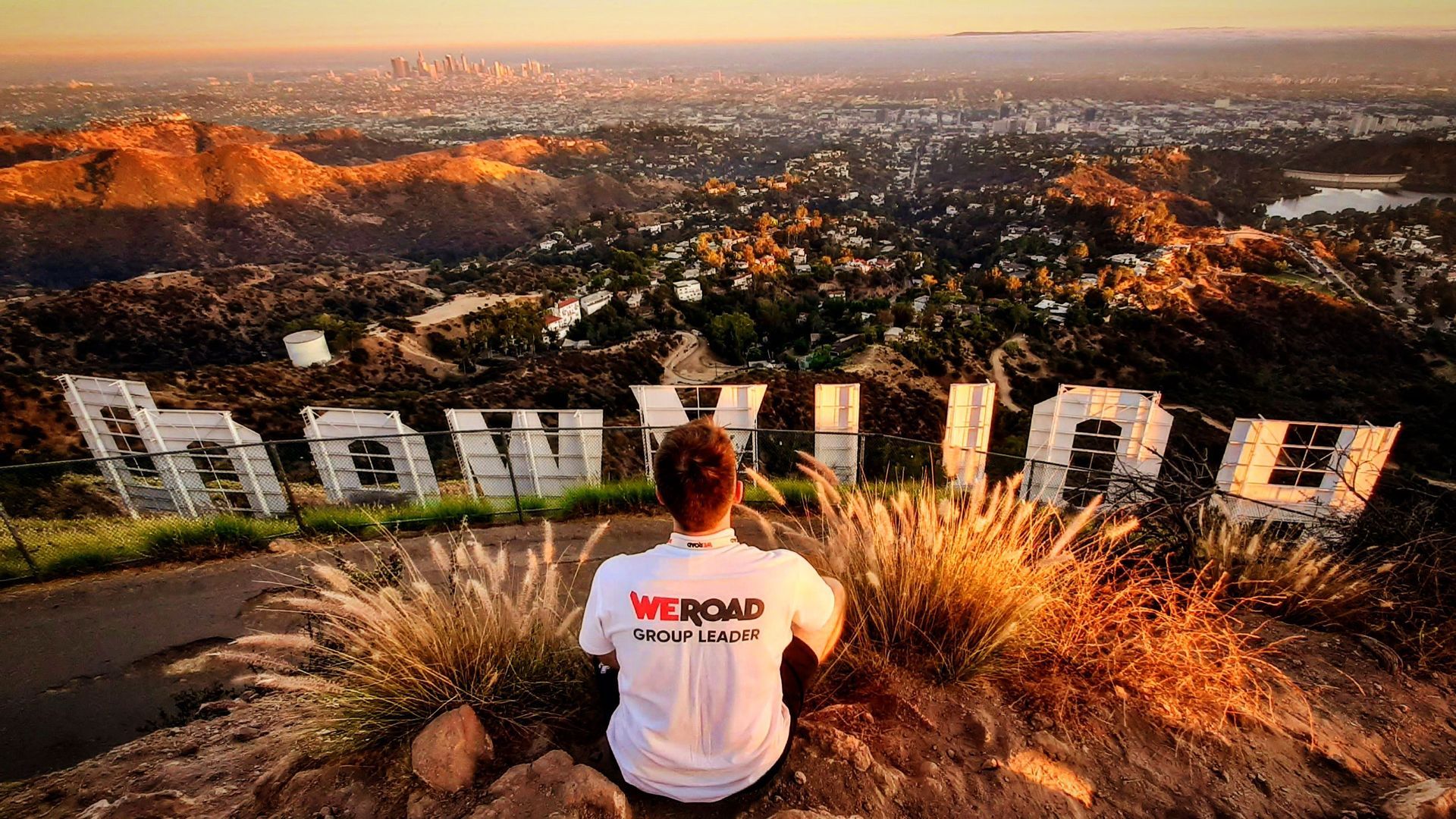 Un Group Leader WeRoad è seduto su una collina dietro la scritta Hollywood, affacciato su una vasta città al tramonto.