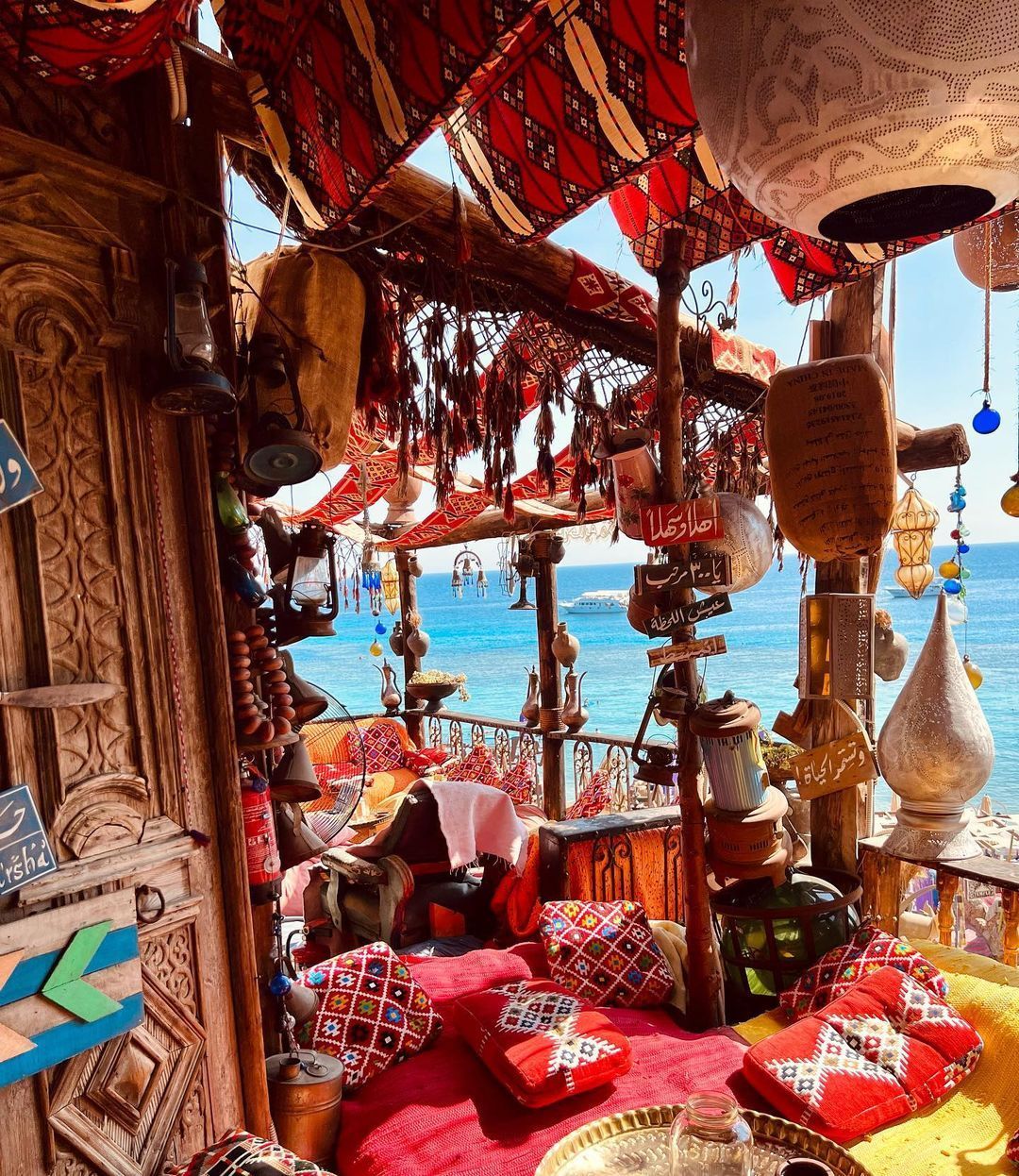 A traditionally decorated balcony with red patterned cushions and hanging lanterns overlooks a bright blue sea on a sunny day.
