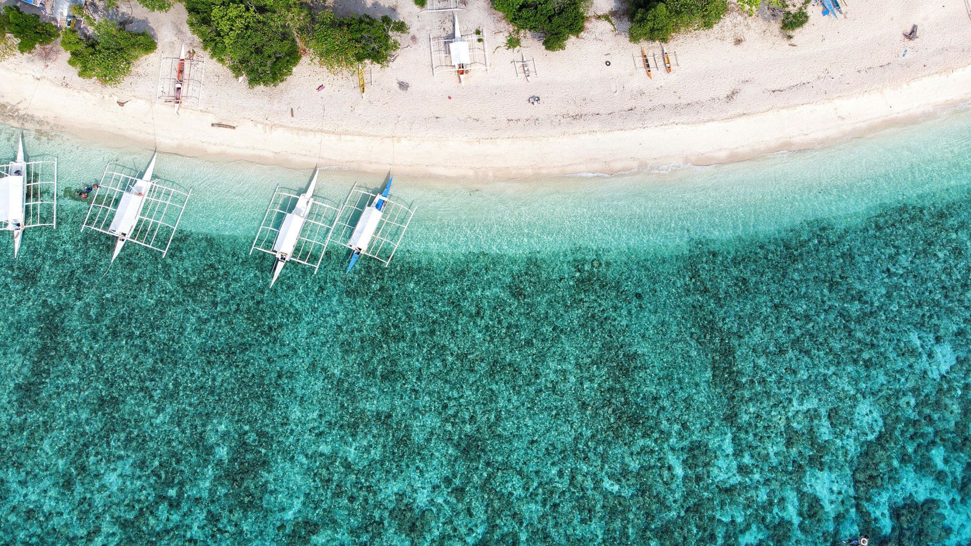 Una vista aerea di barche a bilanciere ormeggiate su una spiaggia di sabbia bianca che costeggia acque turchesi e limpide con una barriera corallina visibile.