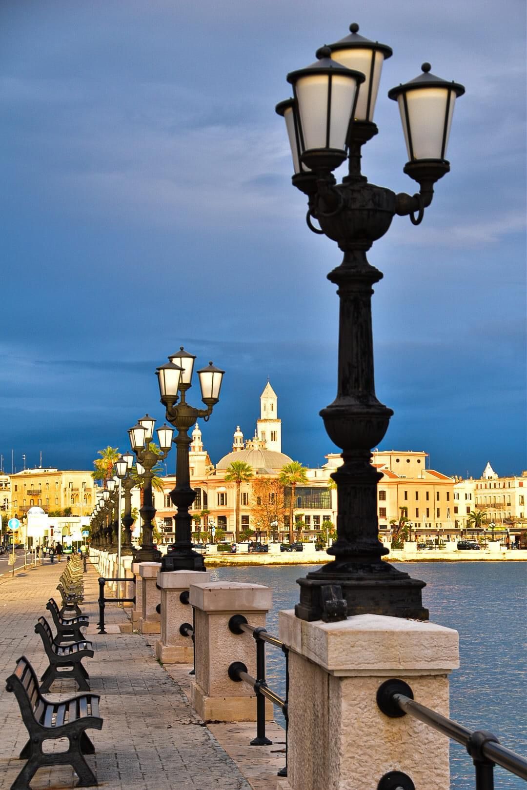 Lampioni neri ornati e panchine costeggiano un lungomare in pietra, con lo skyline di una città costiera sullo sfondo sotto un cielo blu scuro.