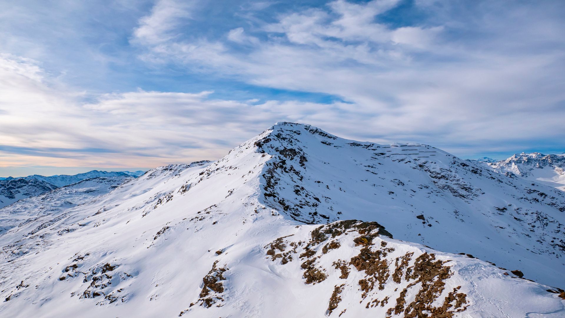 Una cima montuosa innevata con una cresta rocciosa sotto un cielo blu con nuvole sparse.