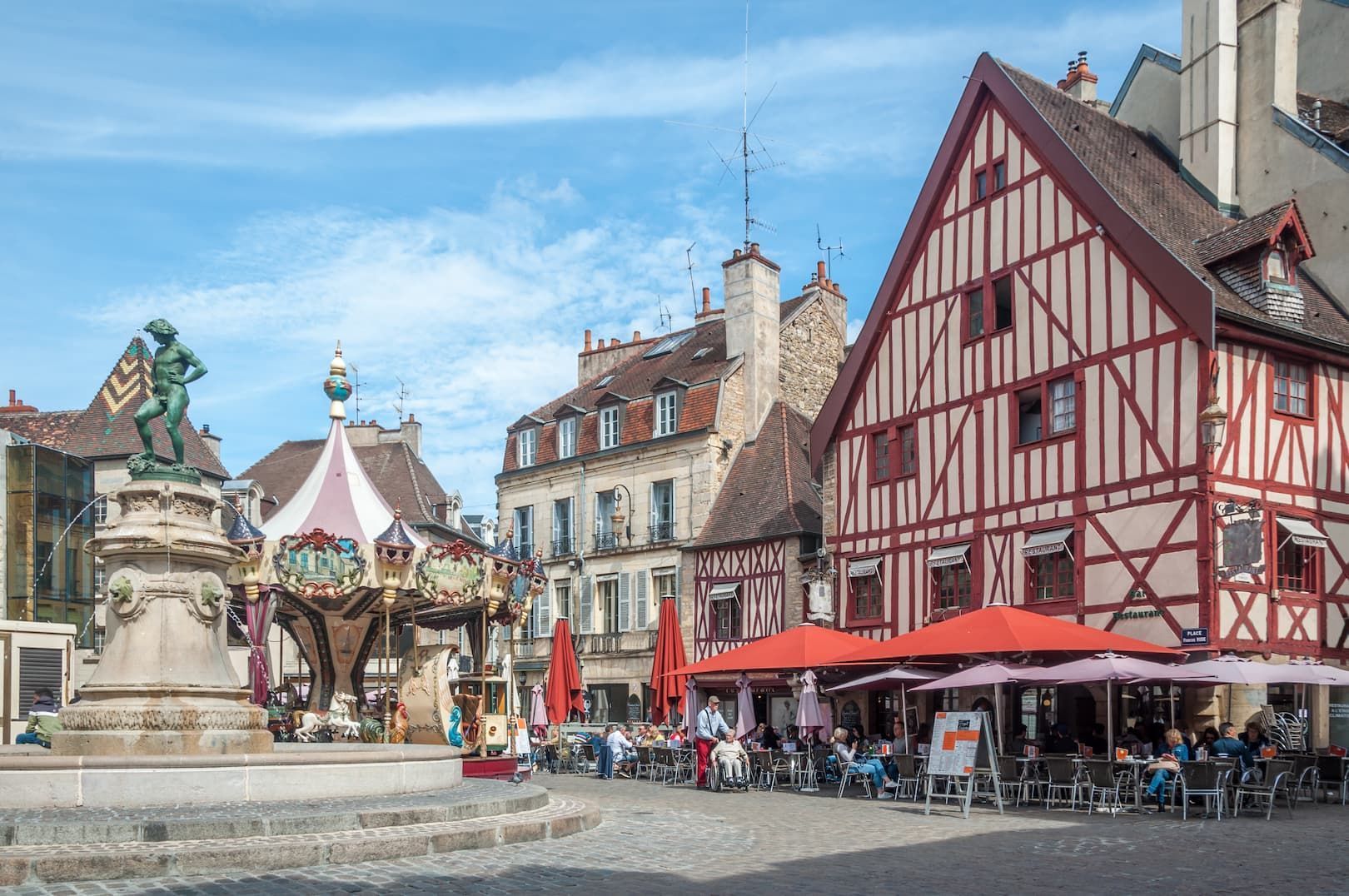 Ein europäischer Stadtplatz mit einem Brunnen, einem Karussell und einem Straßencafé neben traditionellen Fachwerkhäusern.