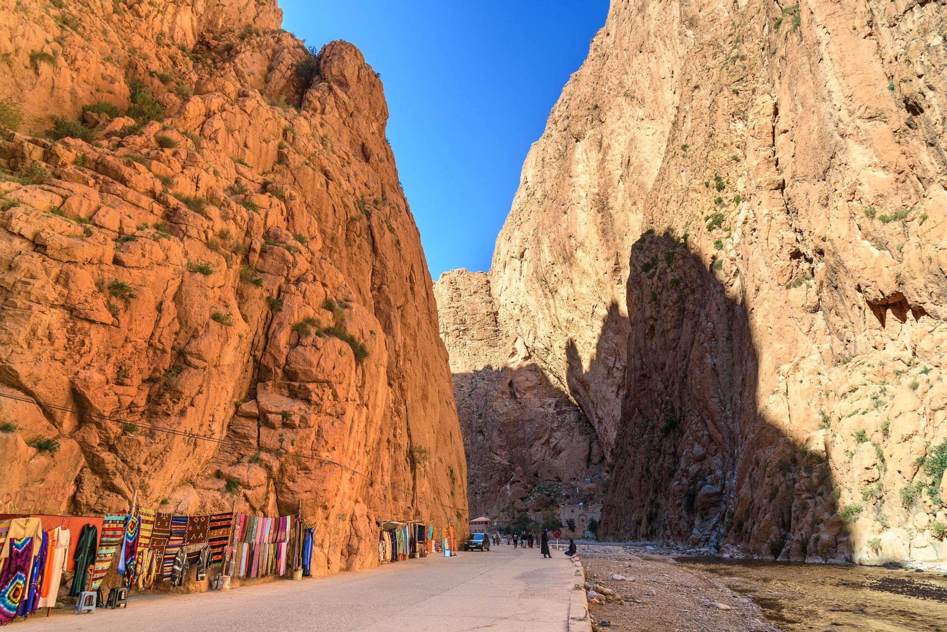 A road lined with market stalls selling colorful textiles winds through a deep canyon with steep, reddish-orange rock walls under a blue sky.