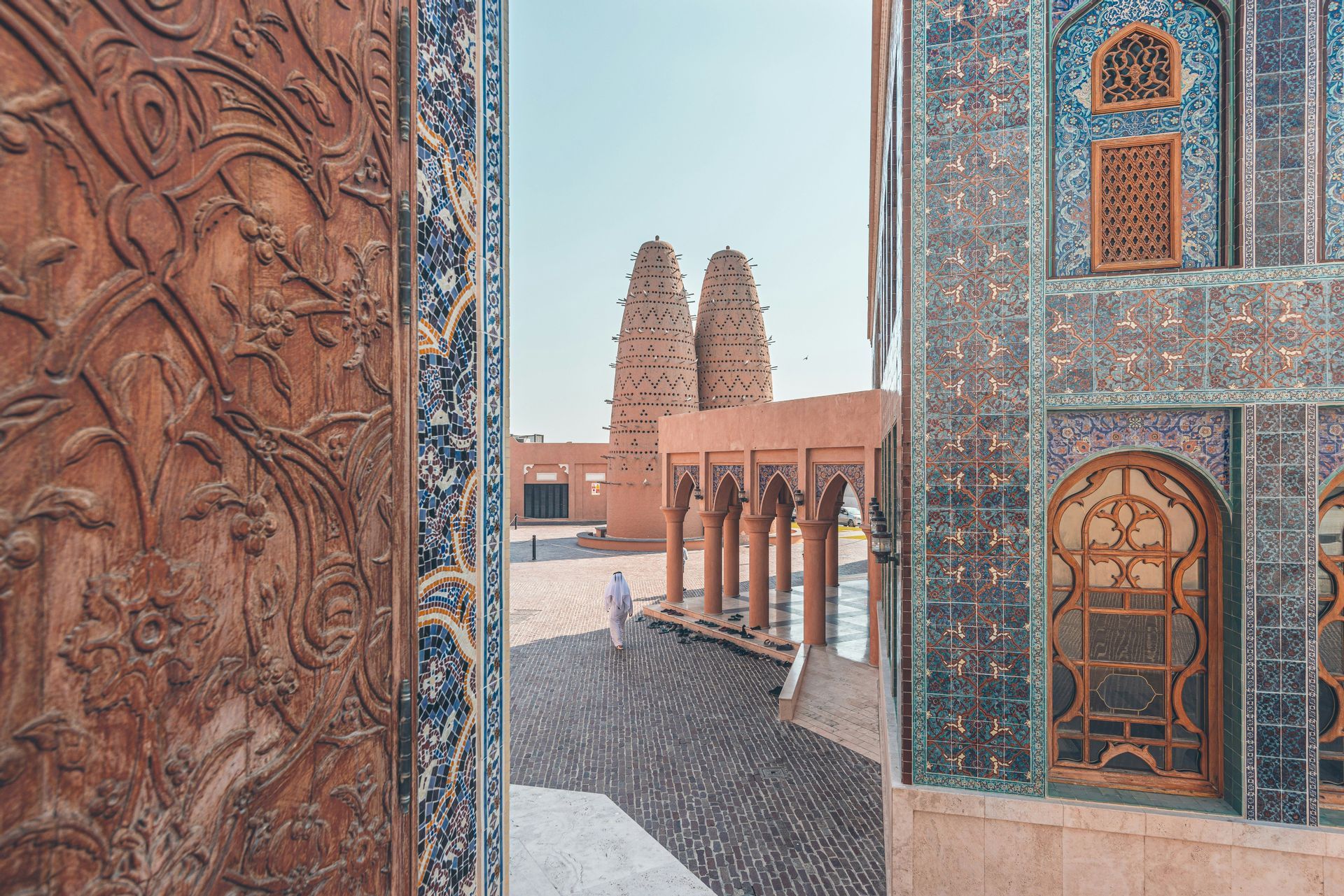 Una persona in abiti bianchi tradizionali cammina in un cortile con torri colombare, vista tra una porta intagliata e un muro a mosaico.