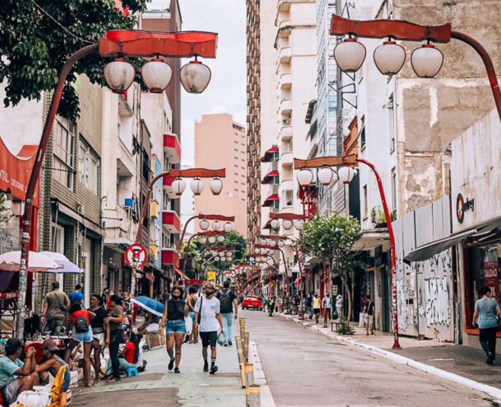 People walk down a busy city street lined with tall buildings and decorated with red, Asian-style lampposts and lanterns.