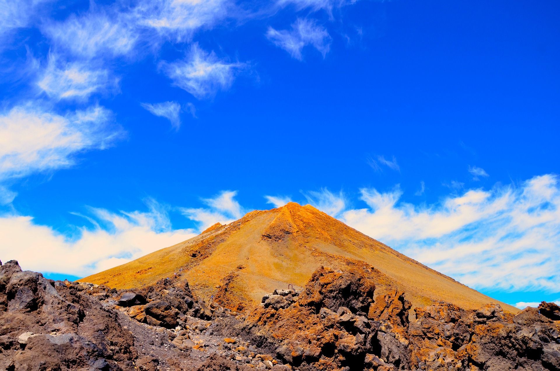 La cima arancione di una montagna vulcanica si erge sopra rocce laviche aspre sotto un cielo azzurro intenso con nuvole sottili.