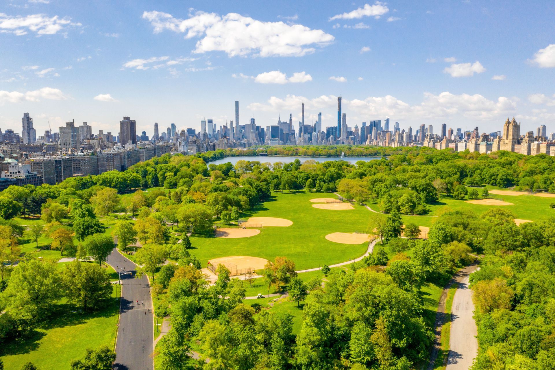 Una veduta aerea di un grande parco urbano con alberi verdi e campi da baseball, con un denso skyline cittadino e un bacino idrico sullo sfondo.