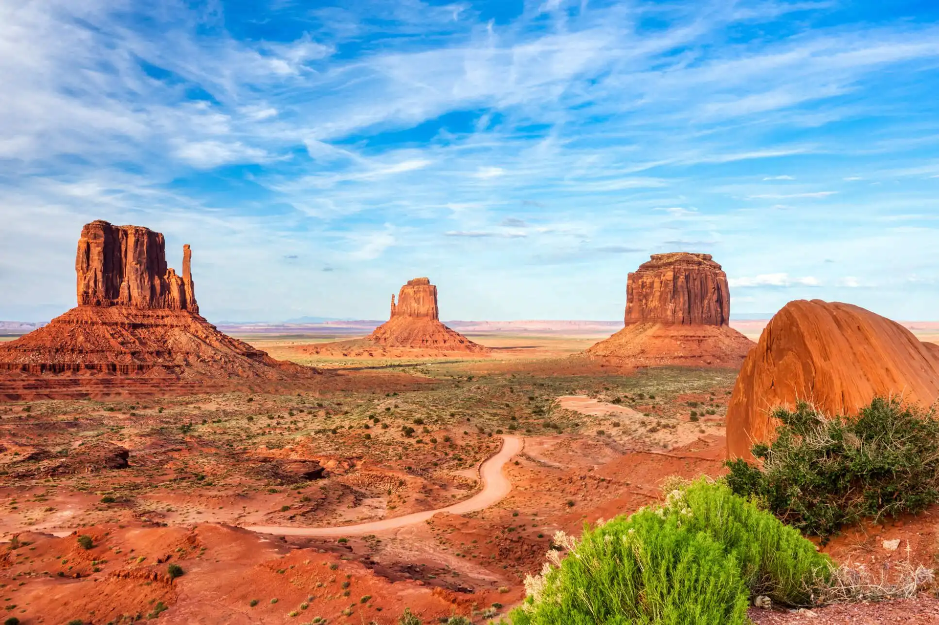 Tre grandi butte di arenaria rossa si ergono in una valle desertica, con una strada sterrata tortuosa e arbusti verdi sotto un cielo blu.