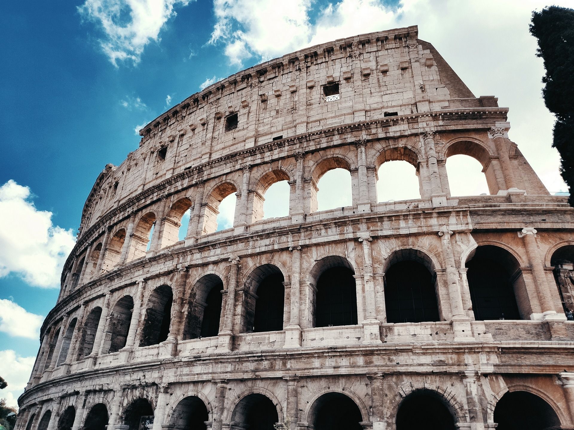 Un'inquadratura dal basso della struttura arcuata in pietra del Colosseo Romano contro un cielo azzurro con nuvole bianche.