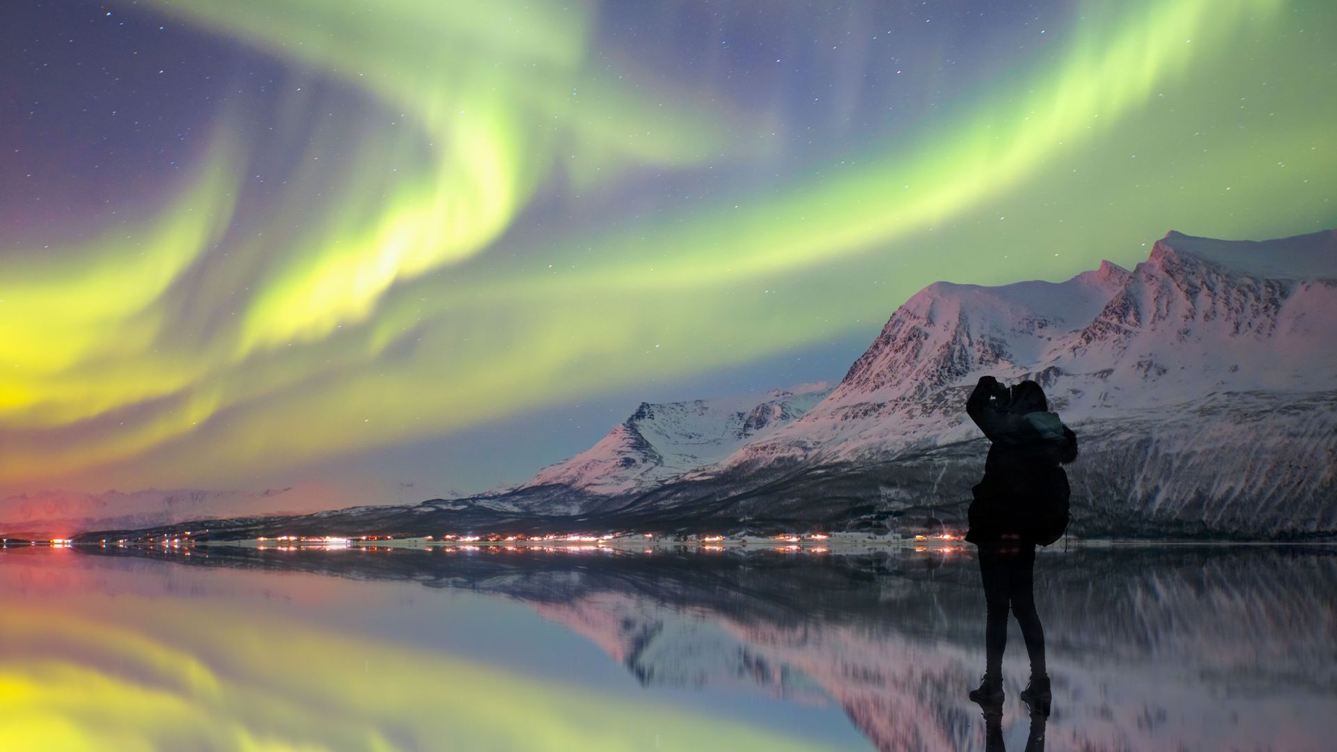 A silhouetted person photographs the green aurora borealis over snow-covered mountains, reflected in calm water at night.