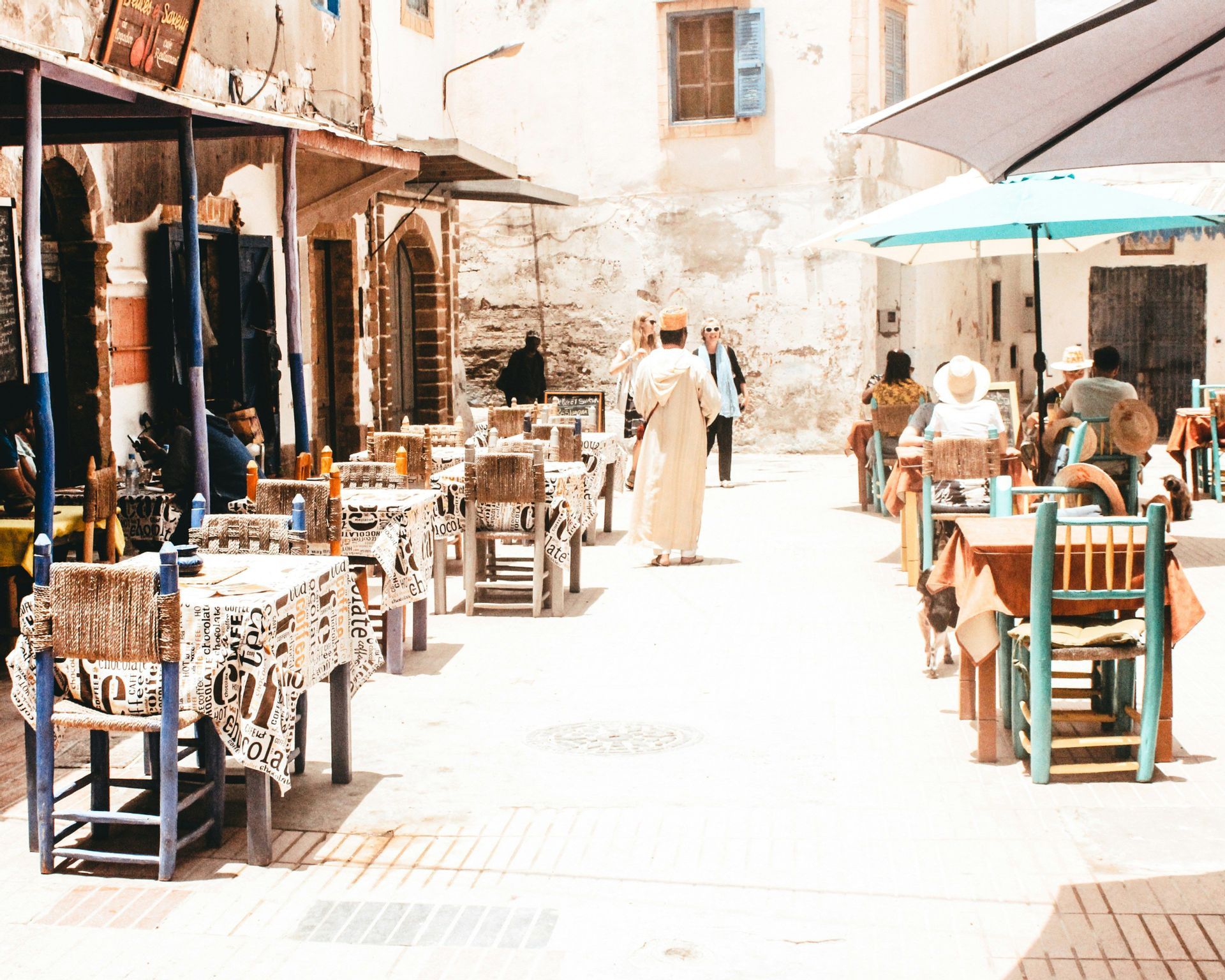 Un caffè all'aperto con tavoli e sedie disposti in una piazza soleggiata e lastricata in pietra, con persone che mangiano e passeggiano.