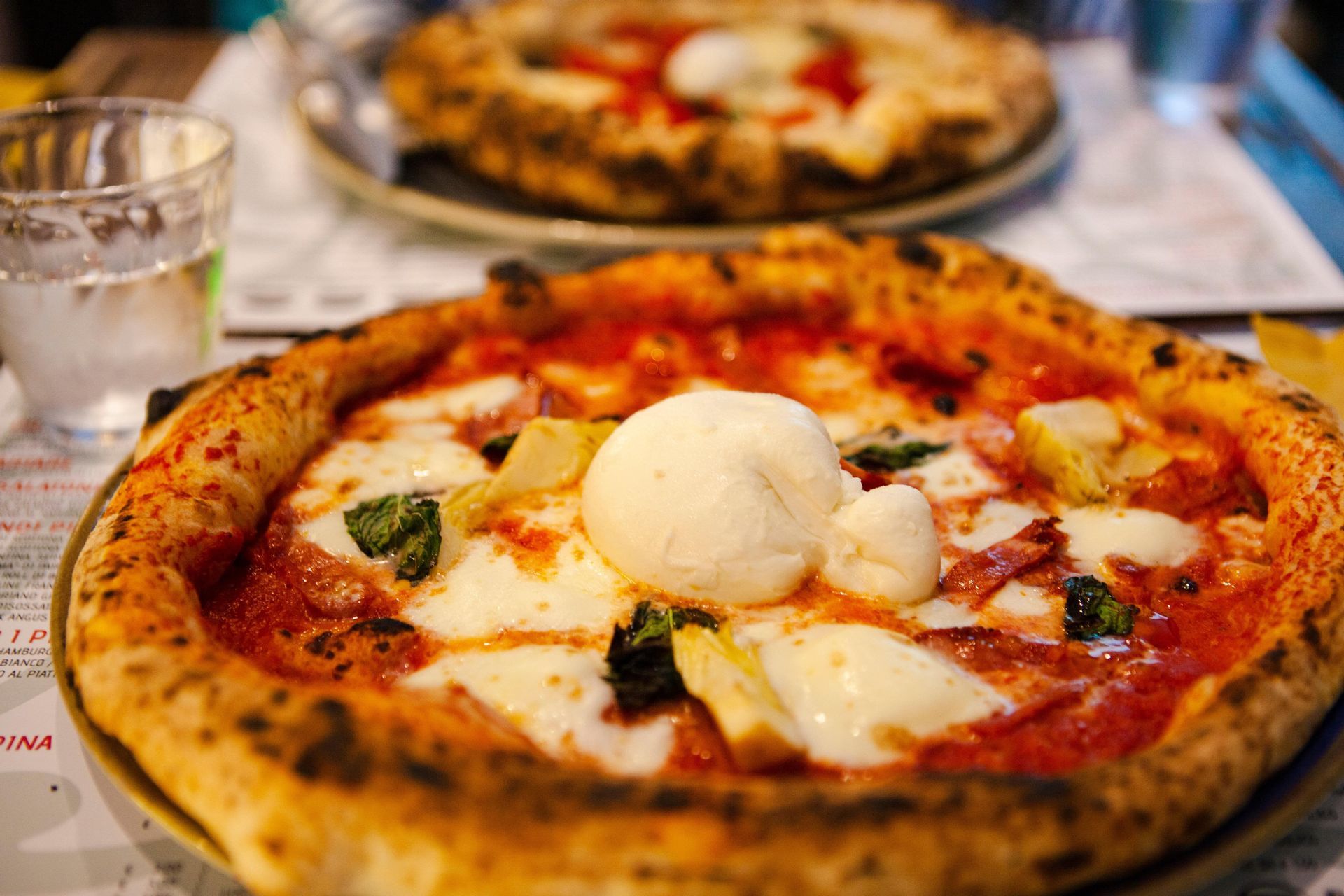 Primo piano di una pizza napoletana con bordo alto, condita con burrata, basilico e carciofi su un tavolo da ristorante.