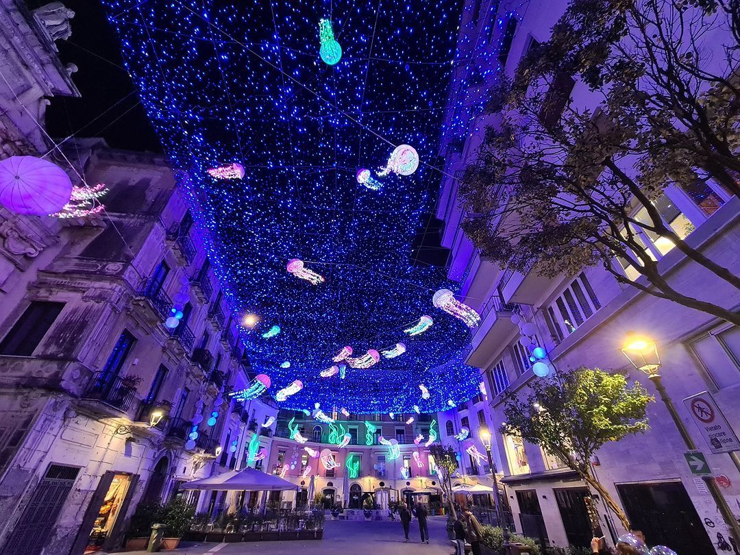 Un soffitto di luci blu e colorate lanterne luminose a forma di medusa pendono sopra una strada cittadina di notte.