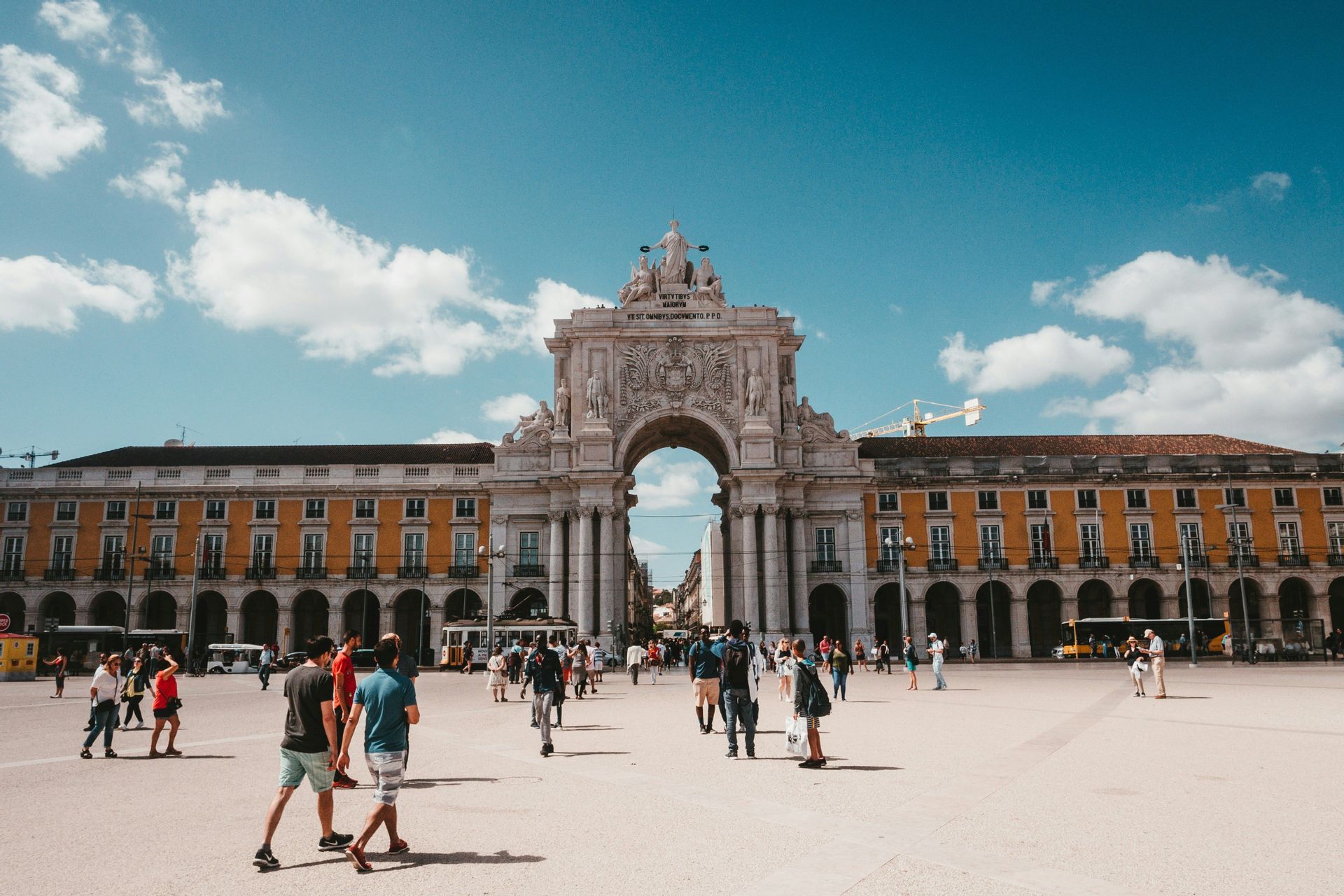Persone che passeggiano in una grande piazza di fronte a un arco trionfale ornato e edifici gialli sotto un cielo azzurro e soleggiato.