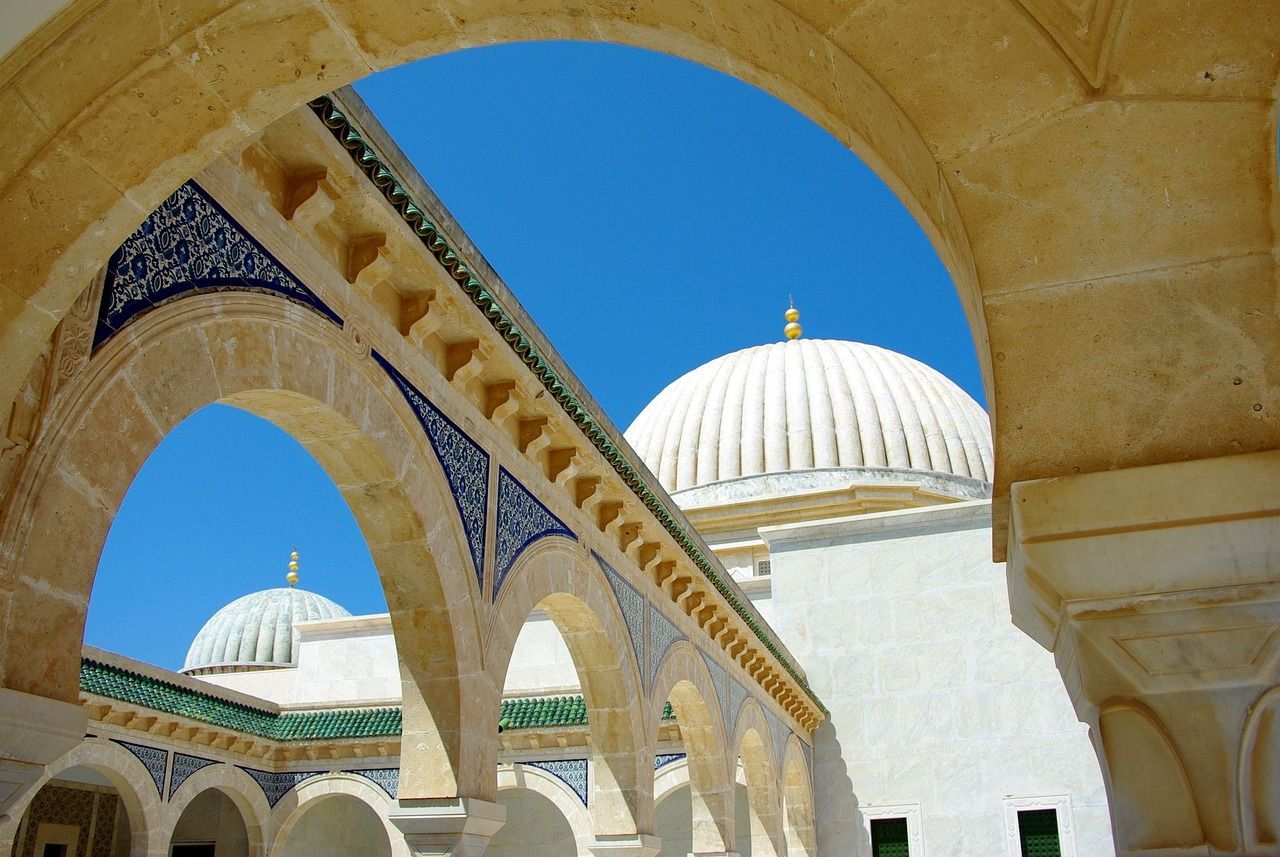 Un arco in pietra color sabbia con dettagli in piastrelle blu incornicia la cupola bianca di una moschea contro un cielo azzurro limpido.