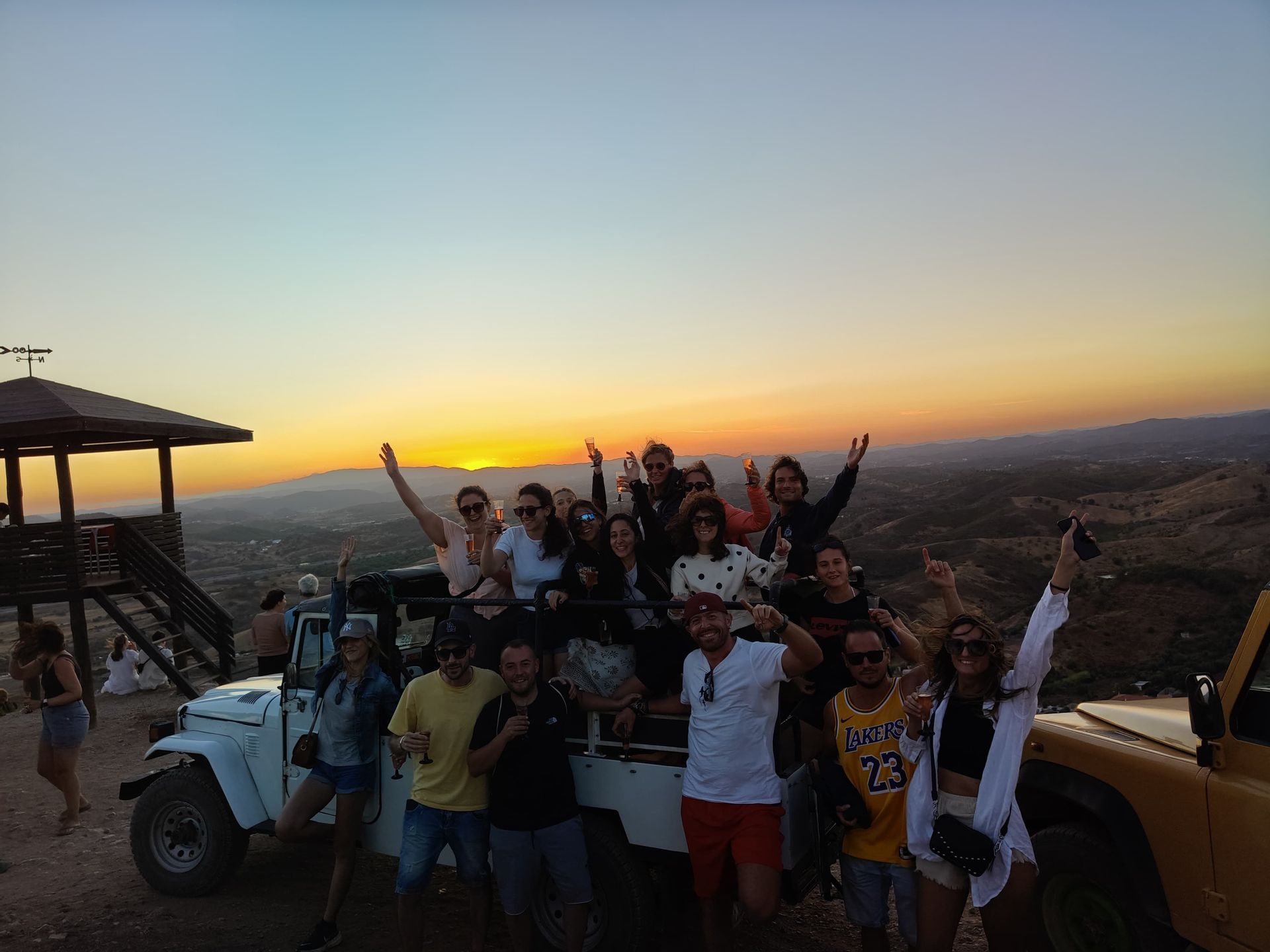 Un gruppo WeRoad in viaggio che posa e festeggia accanto ai fuoristrada su una collina al tramonto.