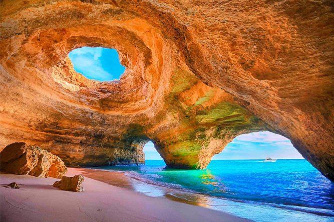 Una spiaggia sabbiosa all'interno di una grotta marina dorata si apre su acque oceaniche turchesi, con un oculo circolare nel soffitto che mostra il cielo blu.