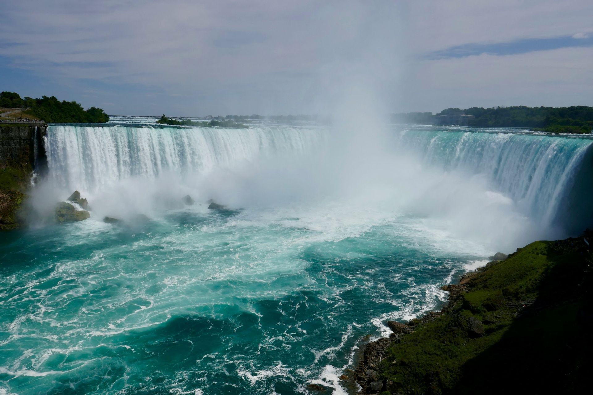 Una cascata ampia, a ferro di cavallo, si getta in un fiume turchese e turbolento, sollevando una grande nuvola di nebbia.