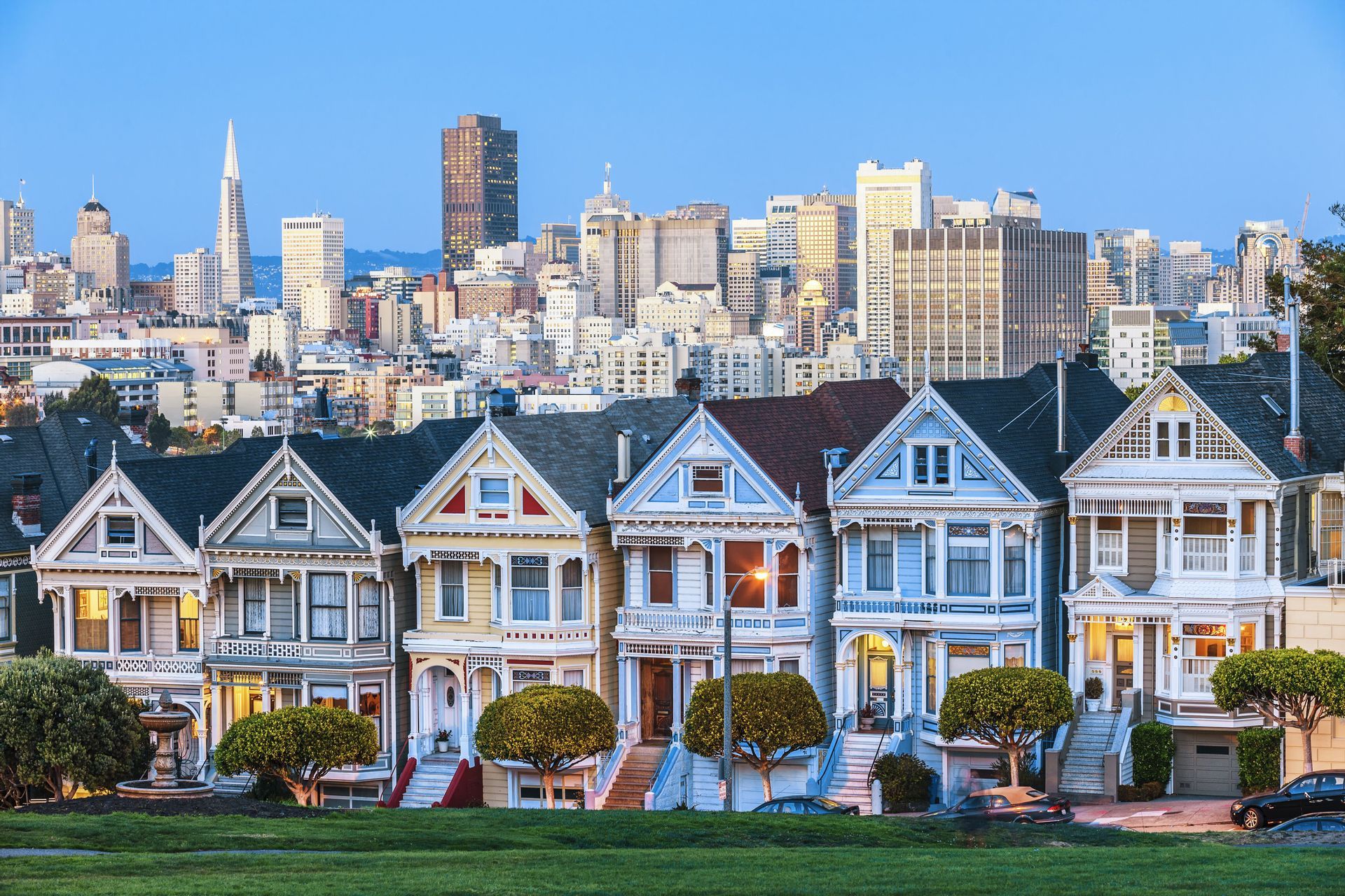 Une rangée de maisons victoriennes colorées se dresse devant un panorama urbain moderne au crépuscule.